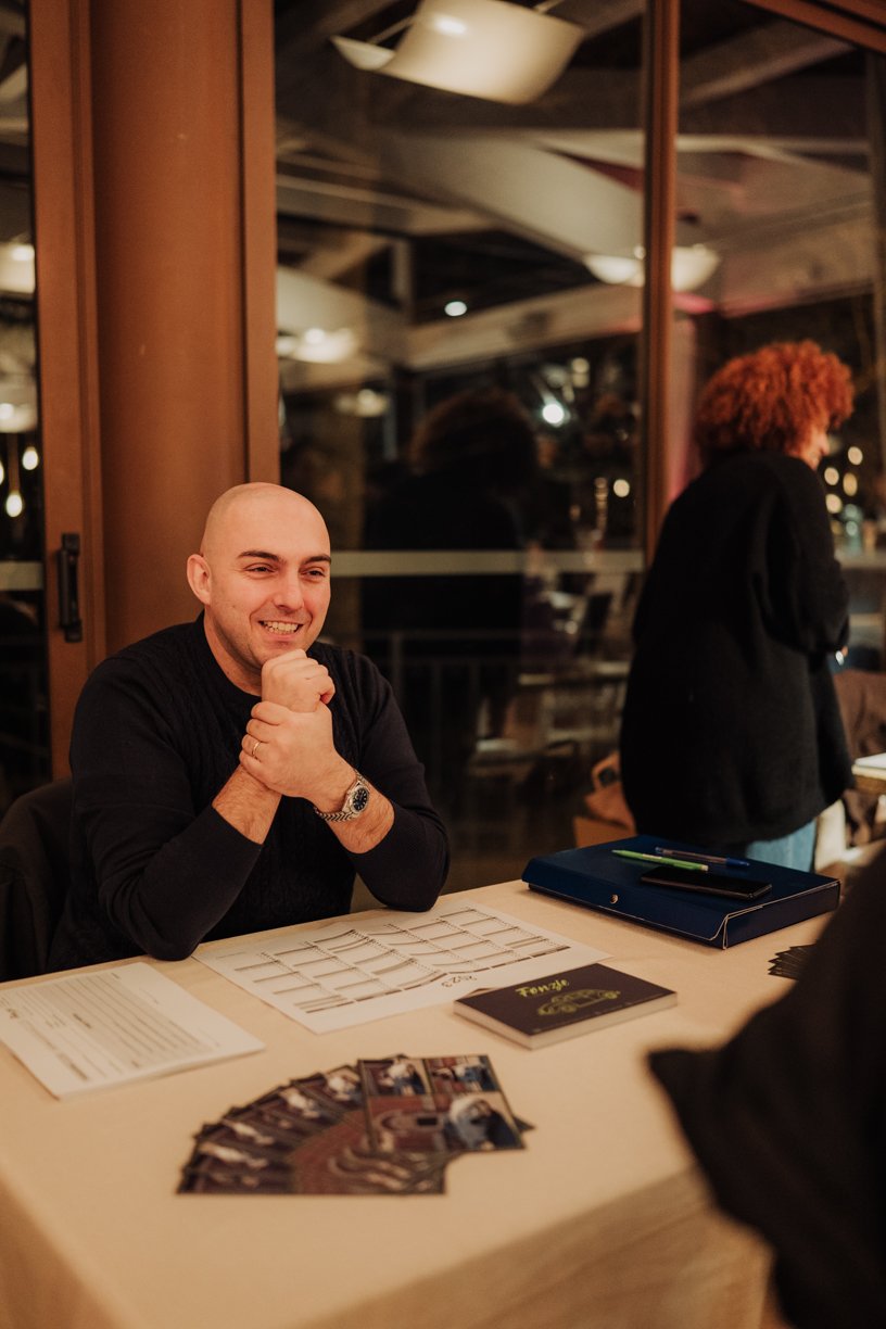 Smiling man at a tabletop with calendar and brochures indoors, with another person standing nearby.
