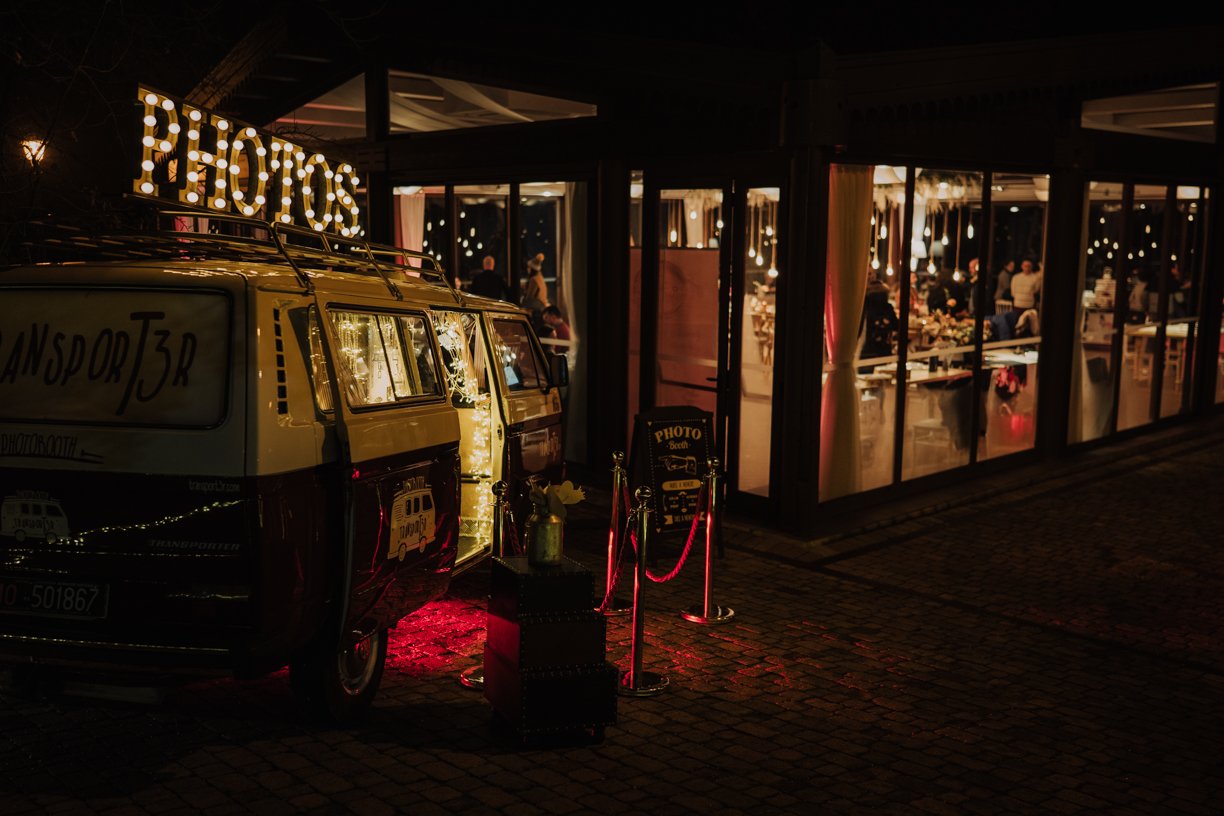 Vintage van converted into a photo booth with a "PHOTOS" sign on top, parked outside a brightly lit event venue with people inside.