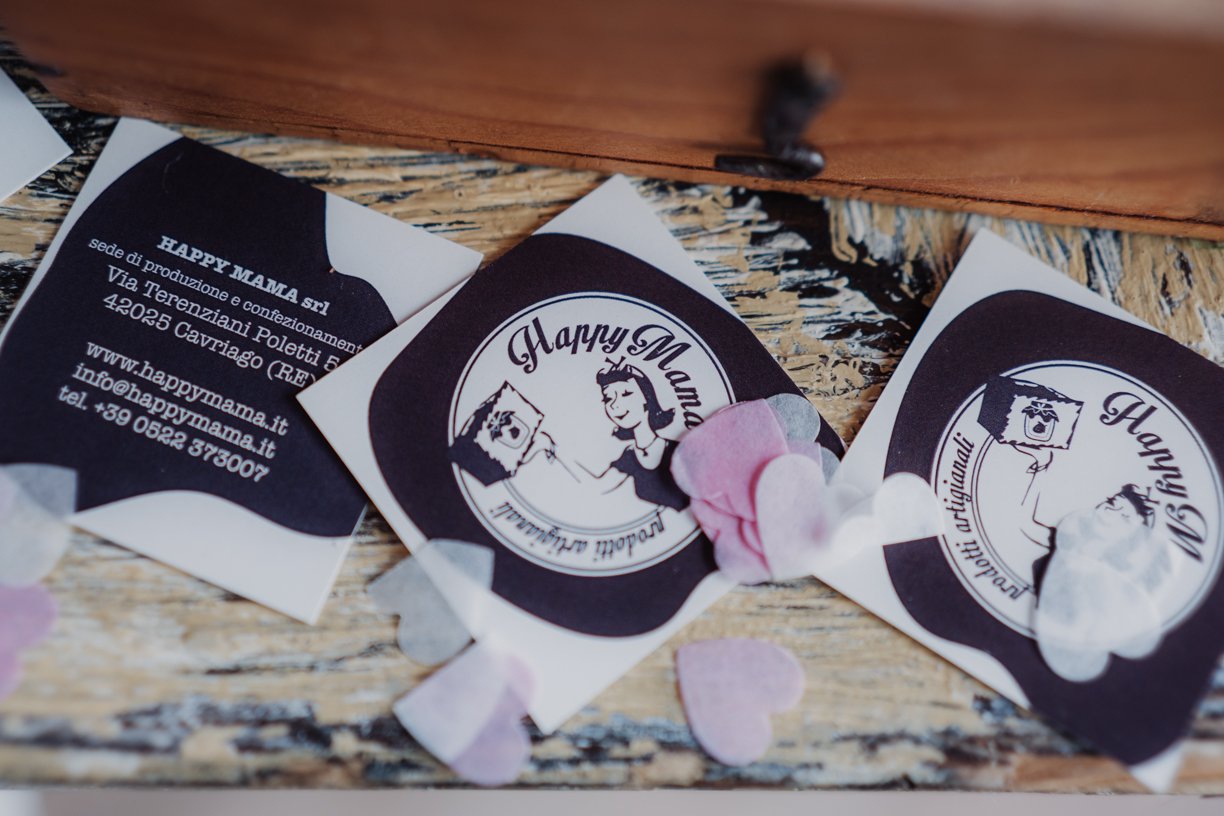 Cards with "Happy Mama" logo and contact information, surrounded by pink and gray heart-shaped confetti on a wooden surface.