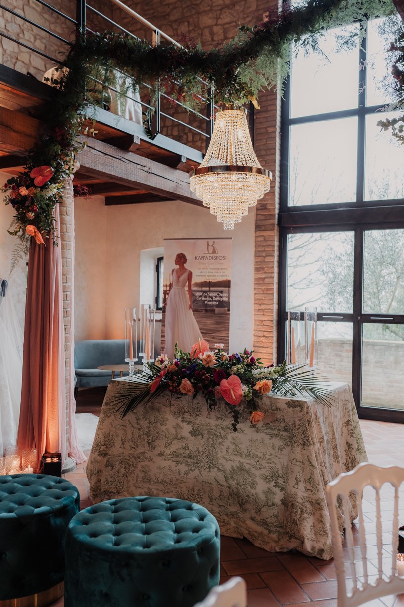 Elegant room setup for a wedding event with a crystal chandelier, floral table decoration, a poster with a bridal gown, and velvet stools.