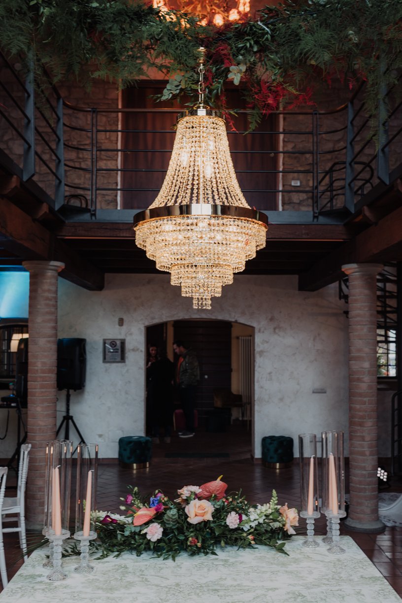 Elegant setting with a large crystal chandelier hanging above a decorated table. The table features floral arrangements and tall candles in glass holders. The room has stone walls, columns, and a balcony with railing. Greenery and lights enhance the