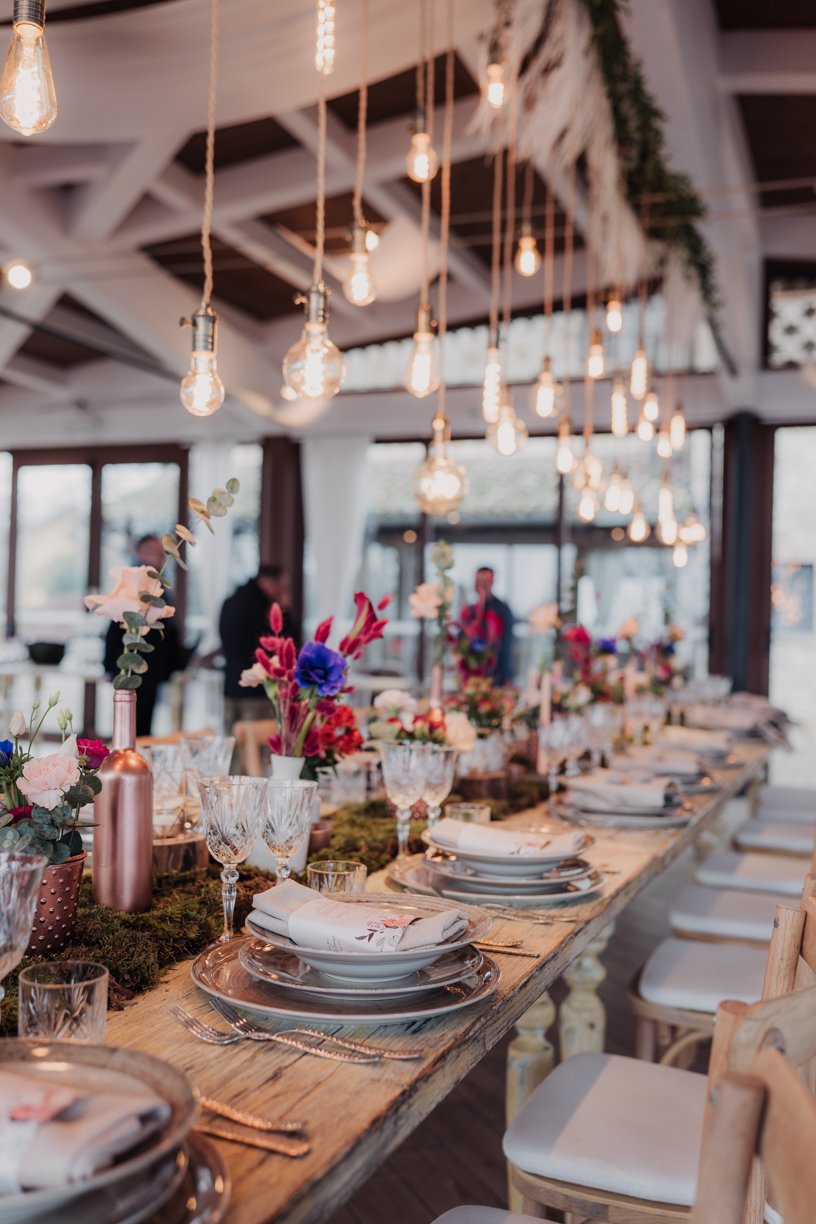 Rustic dining table setup with hanging light bulbs, set for an event with elegant plates, crystal glasses, and vibrant floral centerpieces on a wooden table, surrounded by natural decor.
