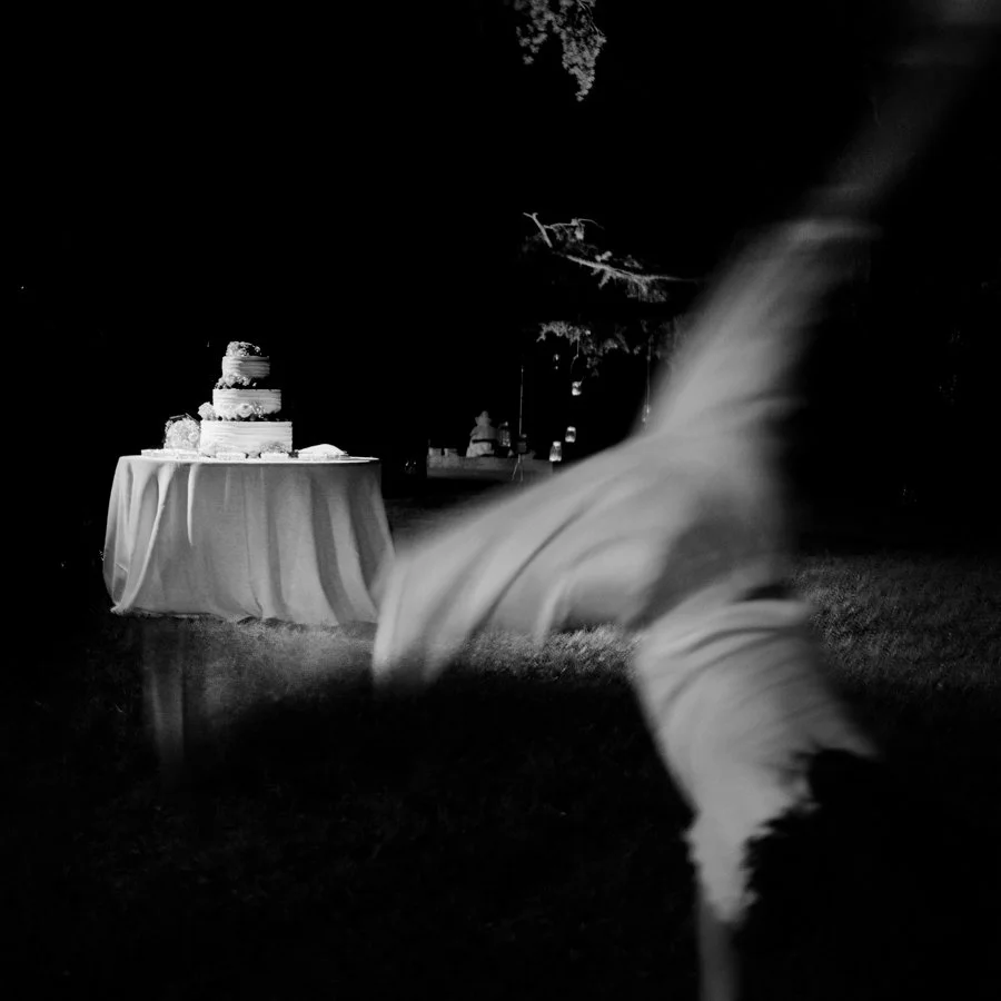 Black and white image of a table with a tiered cake outdoors at night, with a blurred person doing a cartwheel in the foreground.