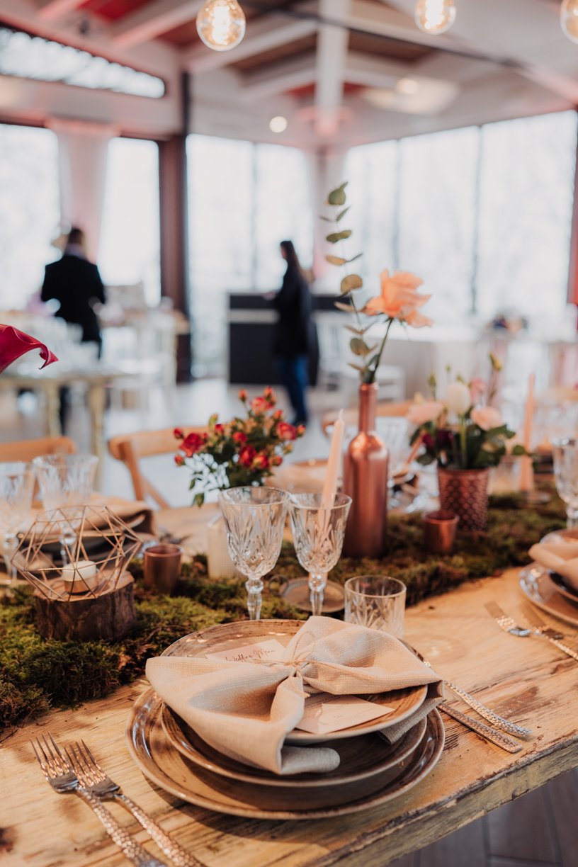 Rustic dining table setup with decorative plates, crystal glassware, and floral centerpieces on a wooden table. Napkins are elegantly placed on the plates, and the setting includes geometric decor and candles. The background shows a person standing i