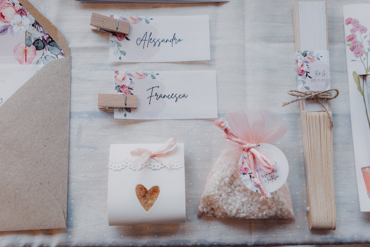 Wedding-themed items including floral place cards clipped with wooden pegs, a small white box with a heart cutout, a pink drawstring bag of rice, a bamboo fan, and a partially visible floral envelope.