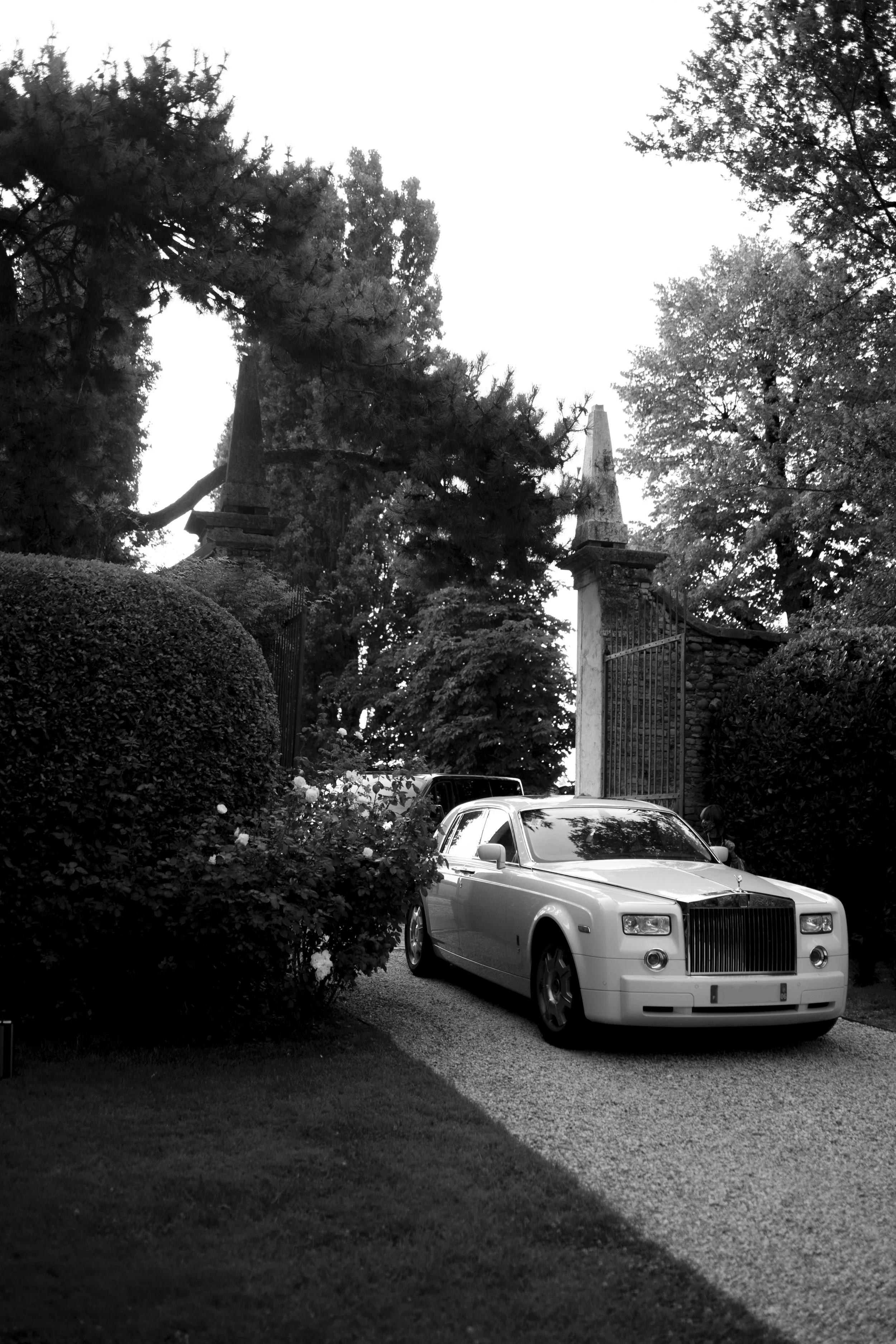 Luxury car parked on gravel road in garden area with trees and hedges.