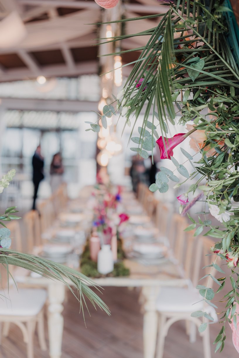 Elegant dining table setup with floral centerpiece and hanging greenery, blurred background with people.
