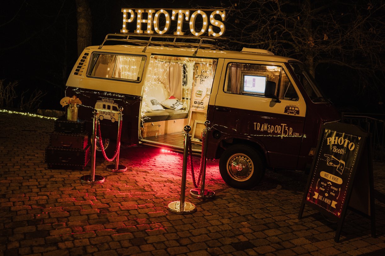 A retro van converted into a photo booth, featuring a "PHOTOS" sign on top, lined with string lights. The van is open, showing a cozy interior with cushions. It is parked on cobblestones with a red carpet and stanchions leading to the entrance. A sig