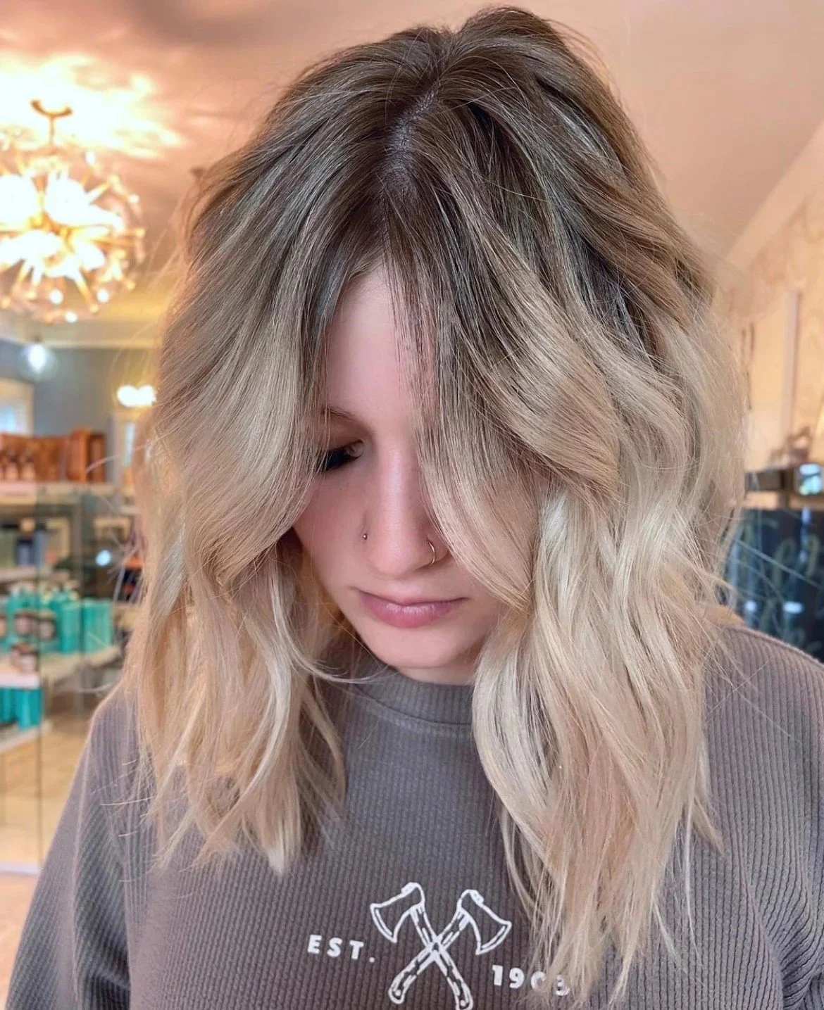 Woman with blonde wavy hair in a salon, wearing a gray sweater with an axe design and the text "EST. 1968."
