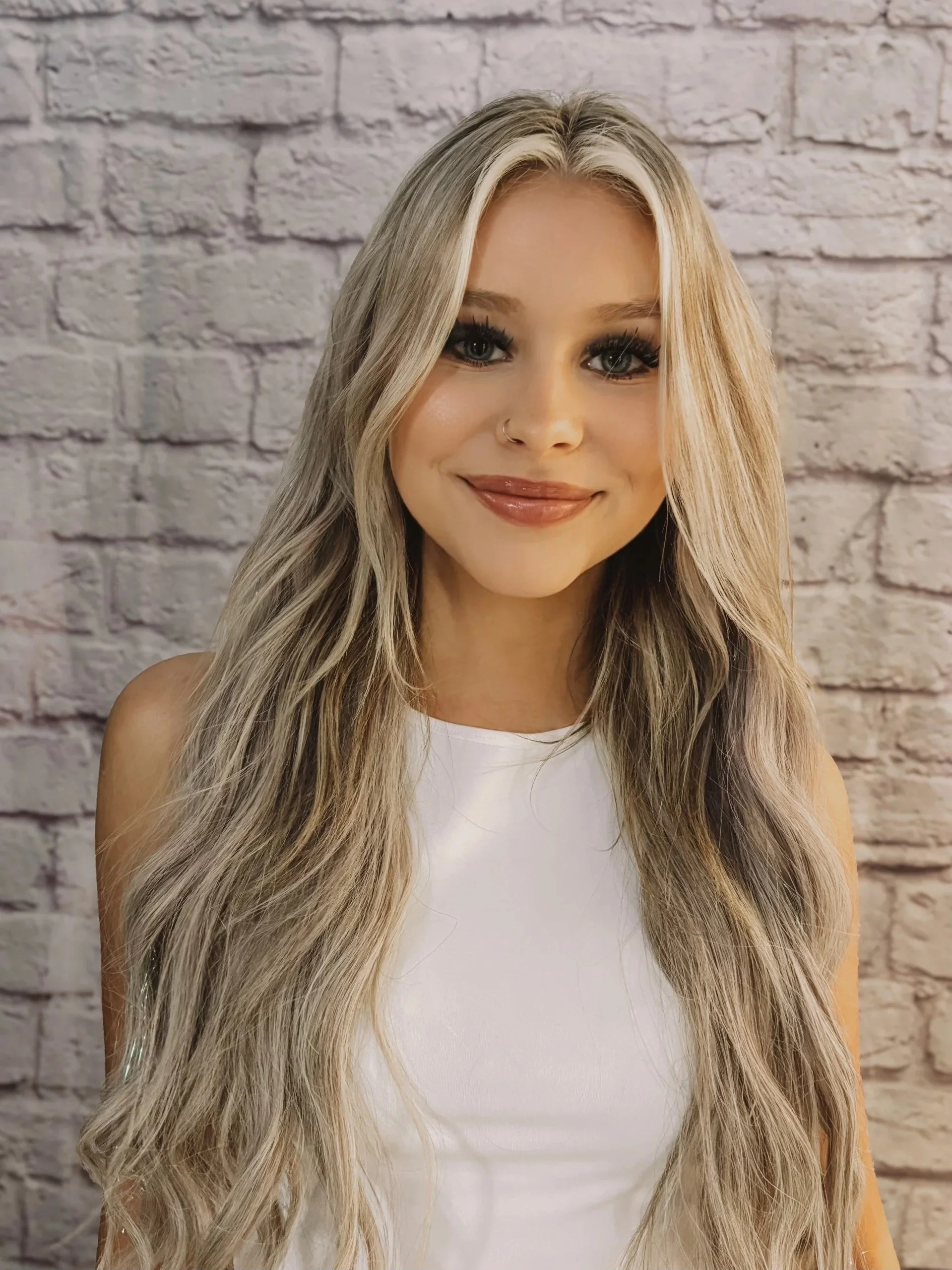 A woman with long, wavy blonde hair, wearing a white top, smiling with makeup and a nose ring, standing in front of a white brick wall.