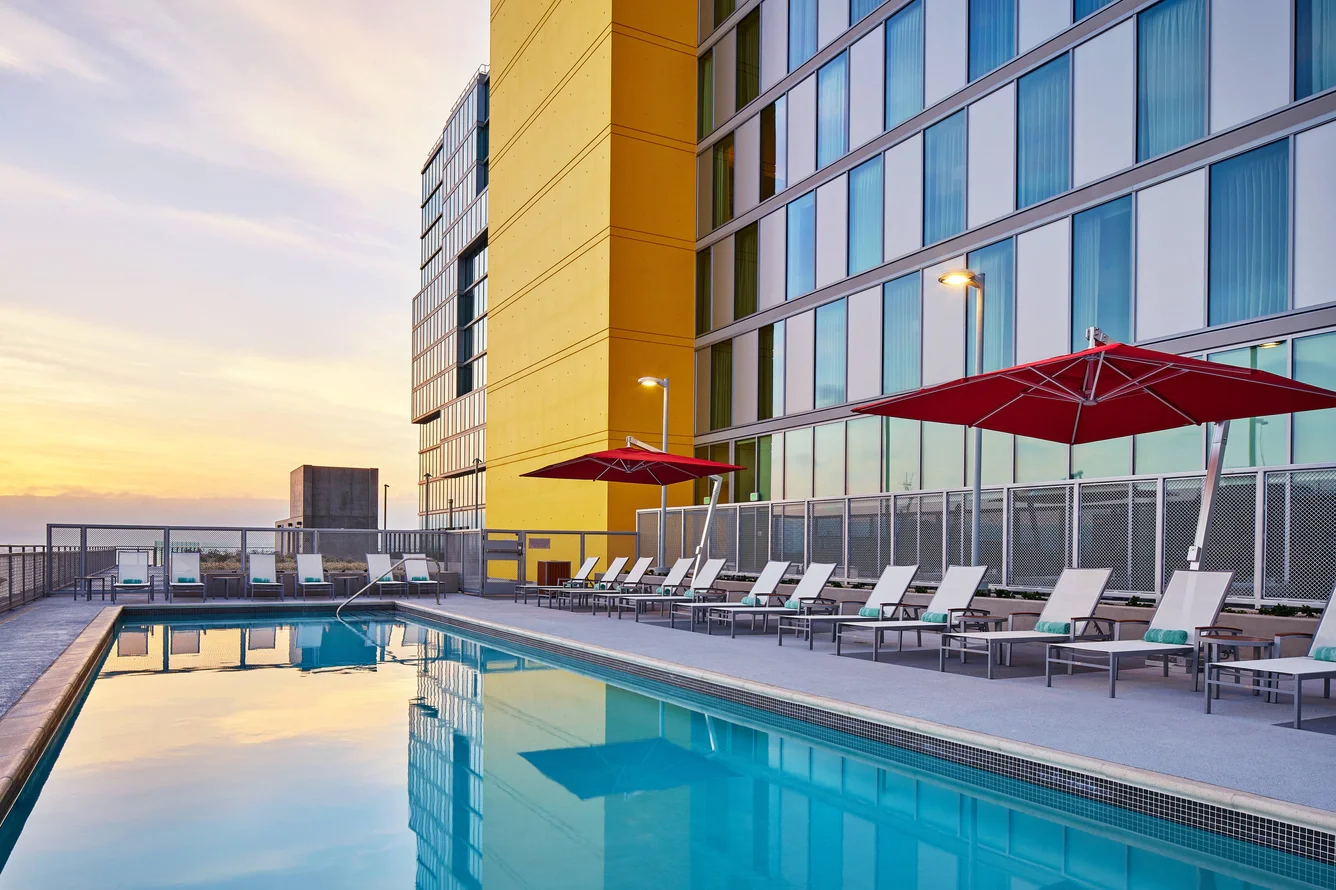 An outdoor rooftop swimming pool at sunset with lounge chairs and red umbrellas, next to a modern glass building.