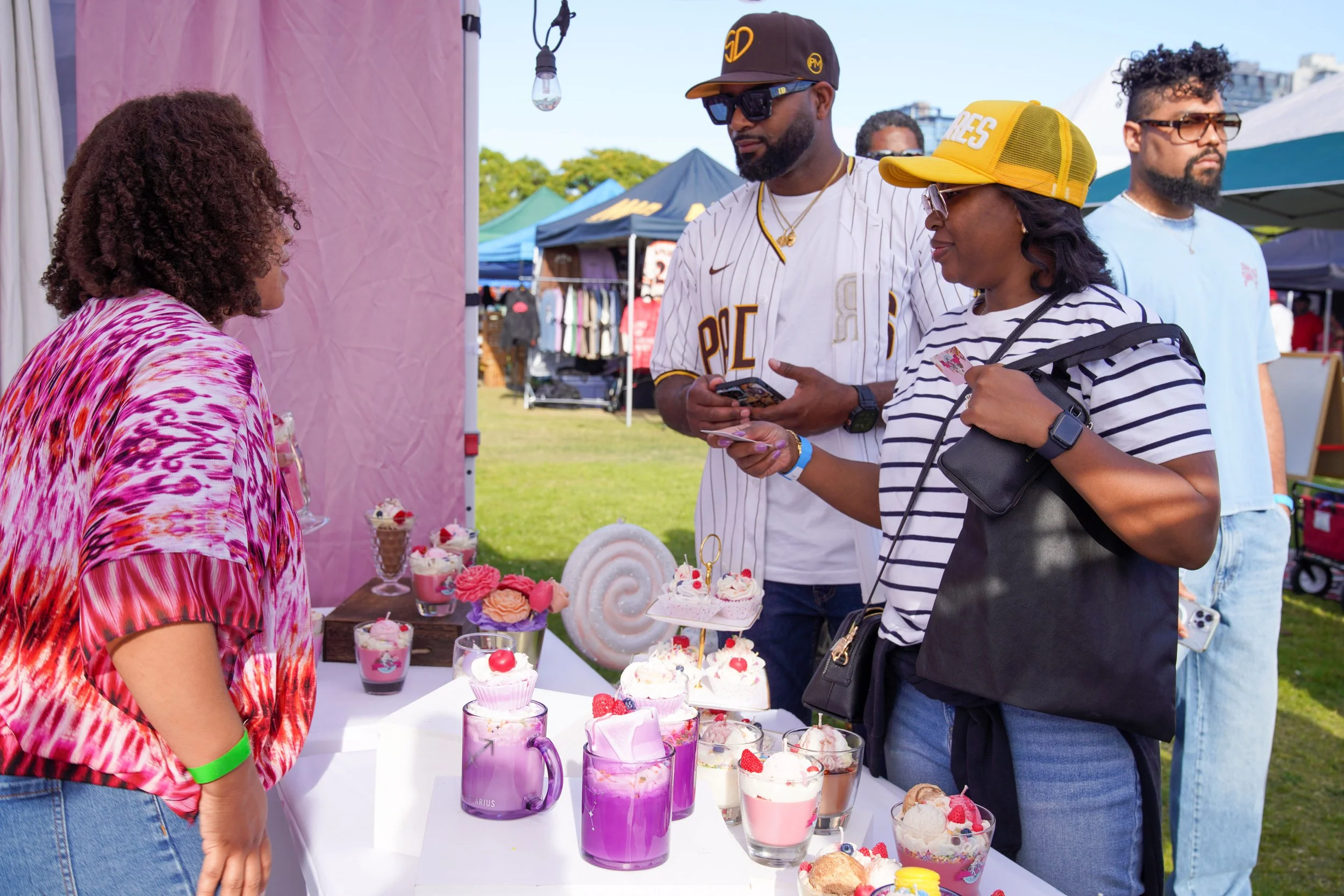 A woman with curly hair wearing a pink and red patterned shirt stands at a table of pink and purple desserts including cupcakes and drinks, talking to a woman in a yellow cap and striped shirt. Two men stand further back at an outdoor event with tents and trees.