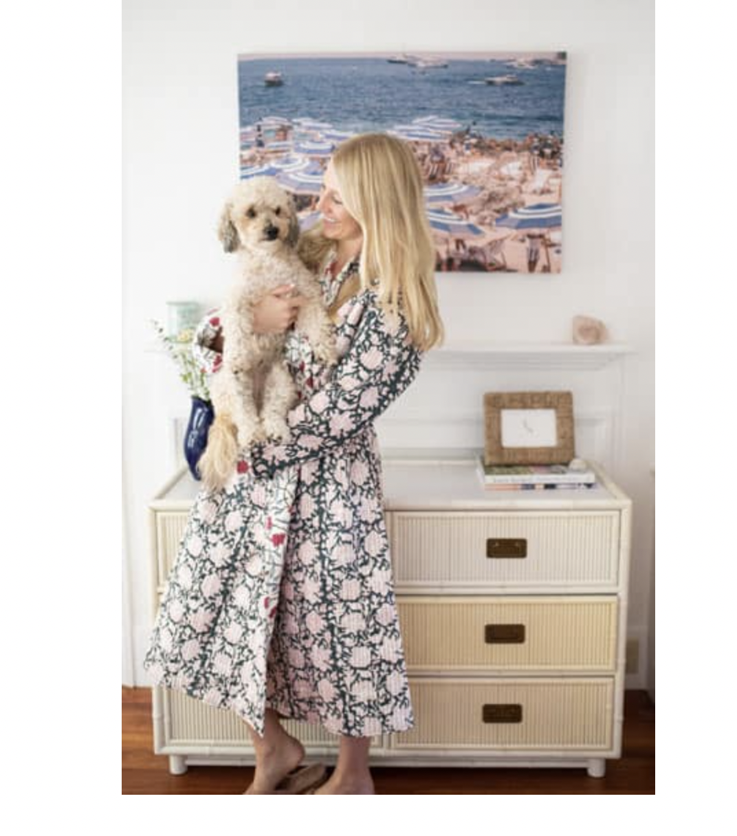 A woman with long blonde hair holding a fluffy dog in an interior room with a white wall and a large seaside painting in the background.