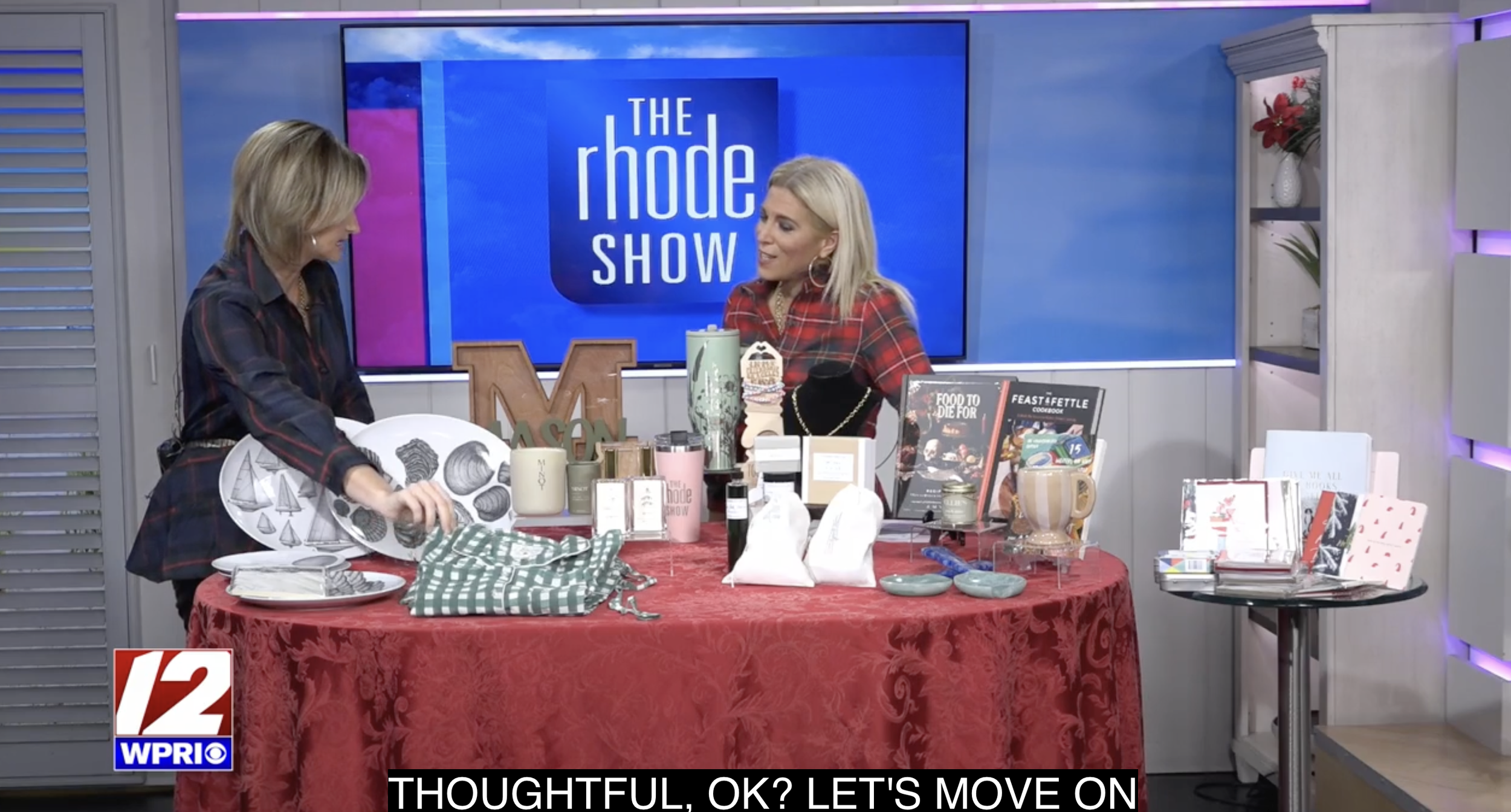 Two women on a television set with a large blue sign reading 'The Rhode Show'. They are discussing various products on a table covered with a red tablecloth, including plates, cups, books, and decorative items.