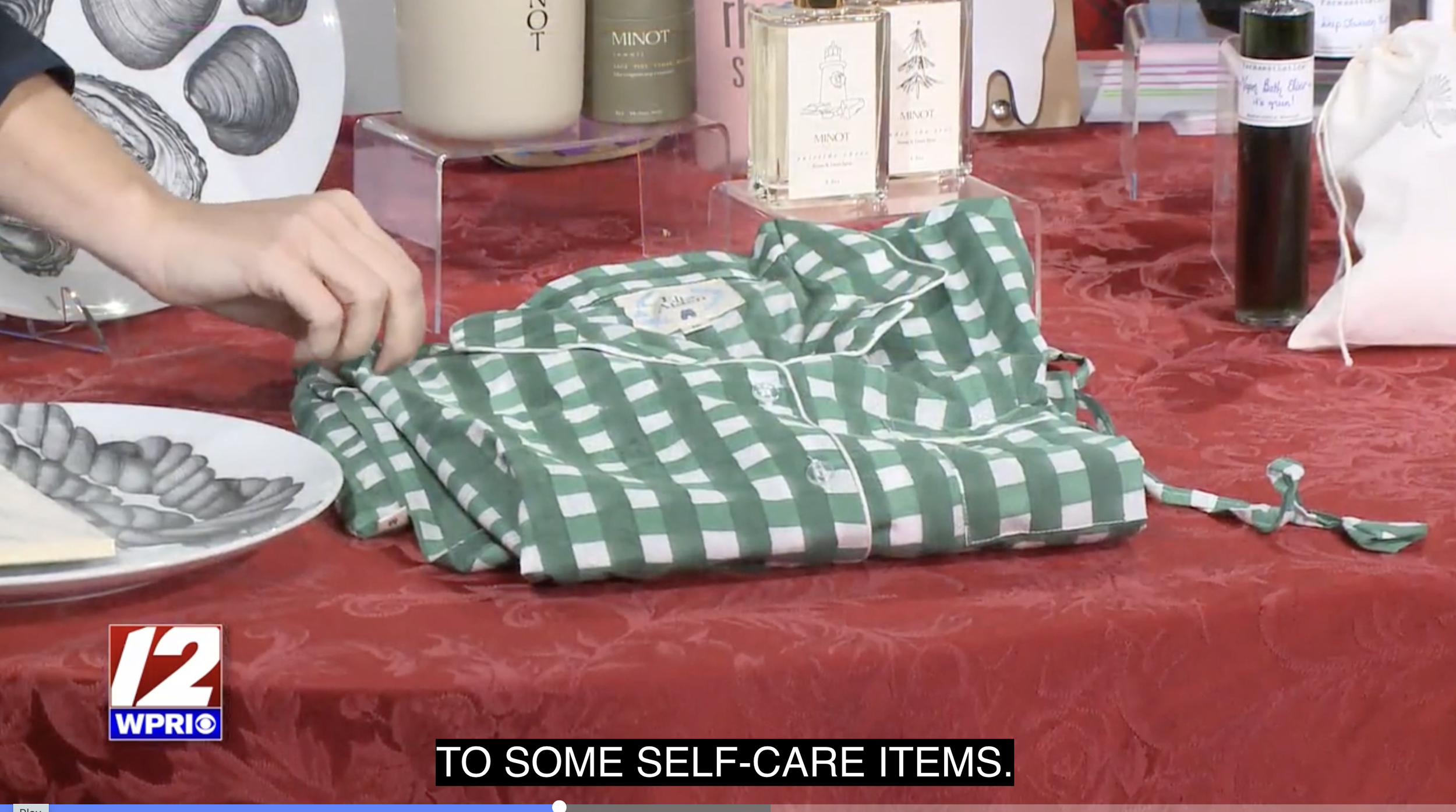 A person's hand is folding a green and white checkered garment on a red table, with various skincare and perfume products displayed in the background.