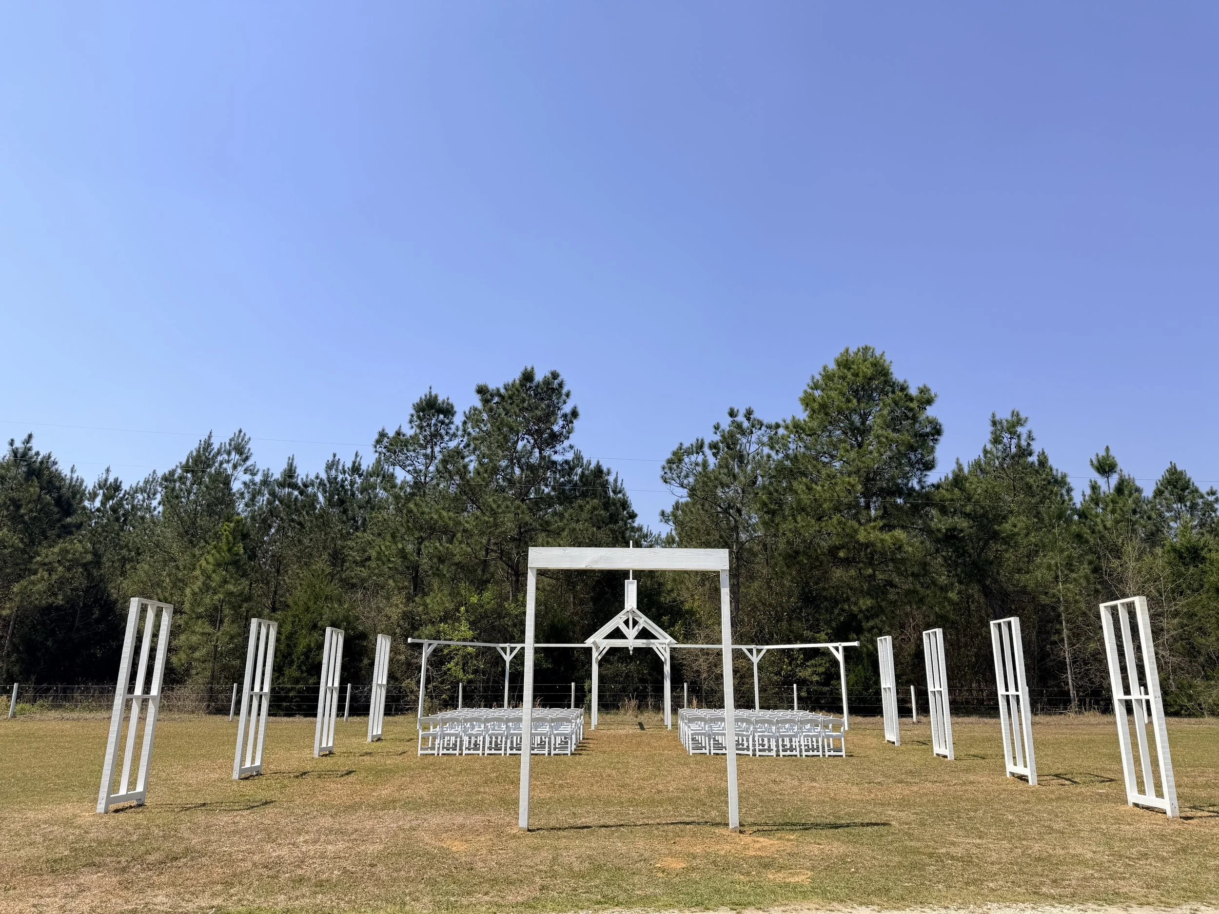 Open Air Chapel ceremony setup with white chairs arranged on a grassy field, white decorative frames, and a canopy, with trees and a clear blue sky in the background.