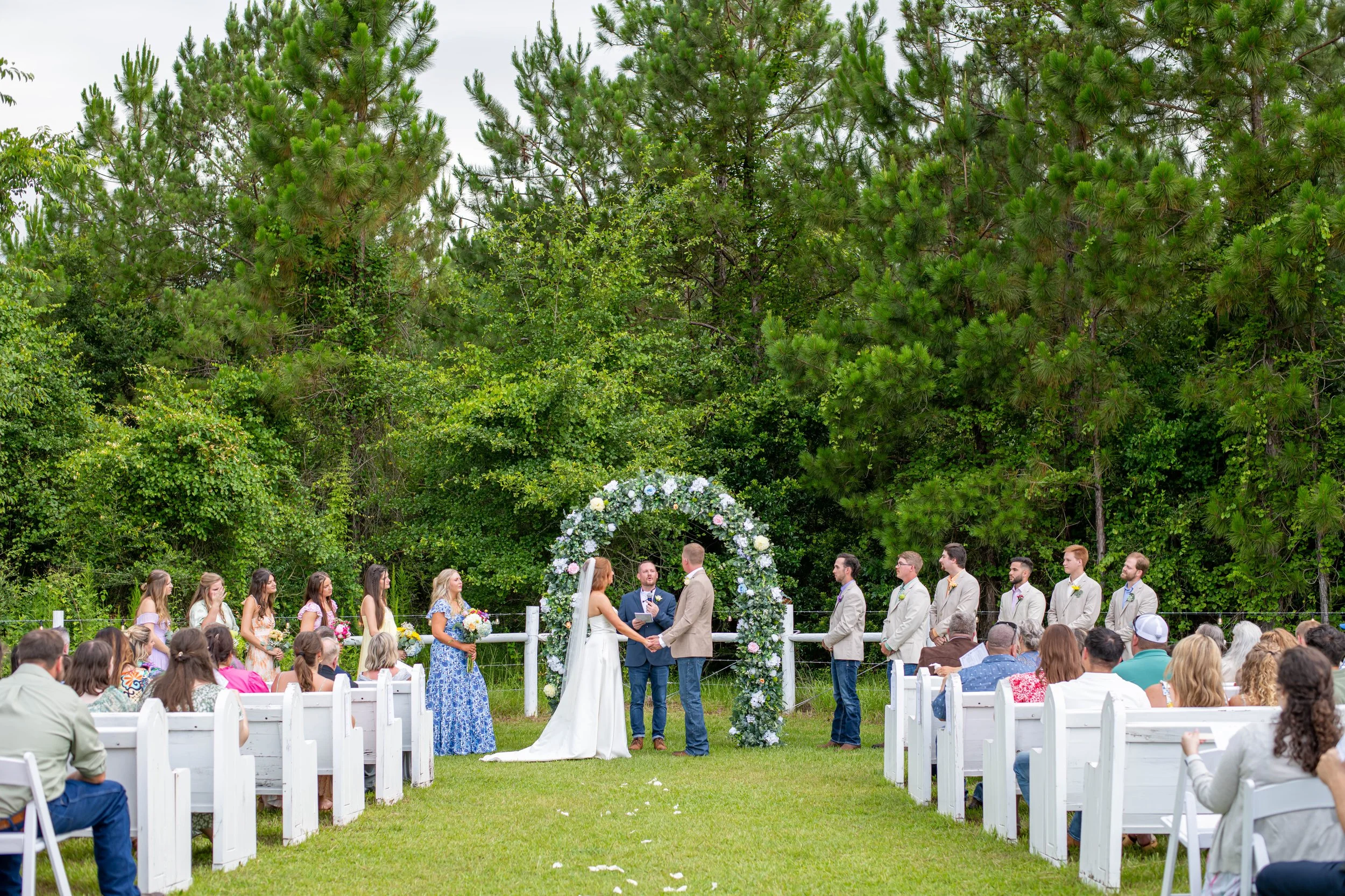 A wedding ceremony outdoors under a leafy green canopy with guests seated on white chairs, the bride and groom standing before an officiant, and a floral arch.