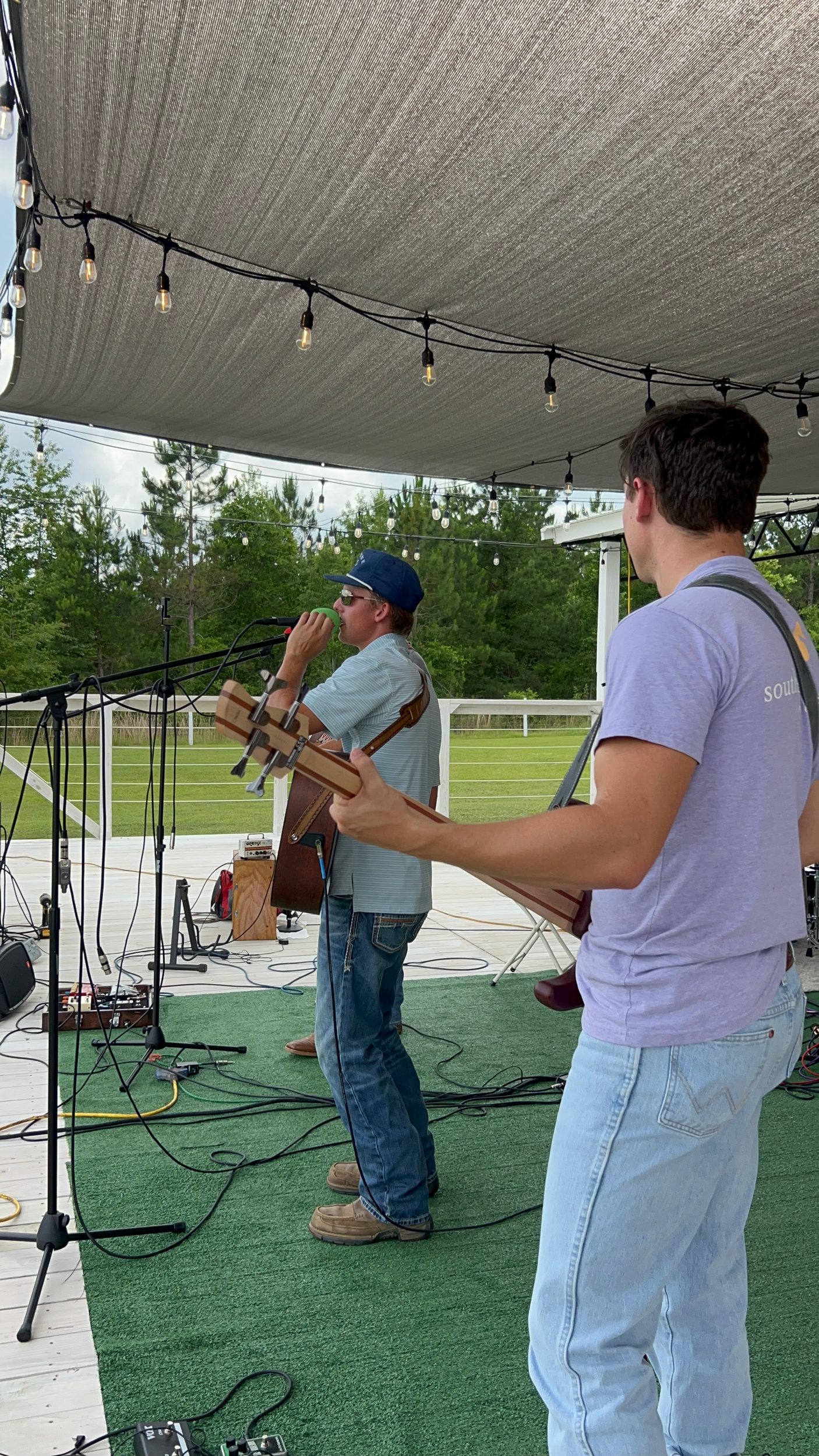 Two people on an outdoor stage with a black canopy and string lights, preparing for a performance, with musical equipment and a green field in the background.