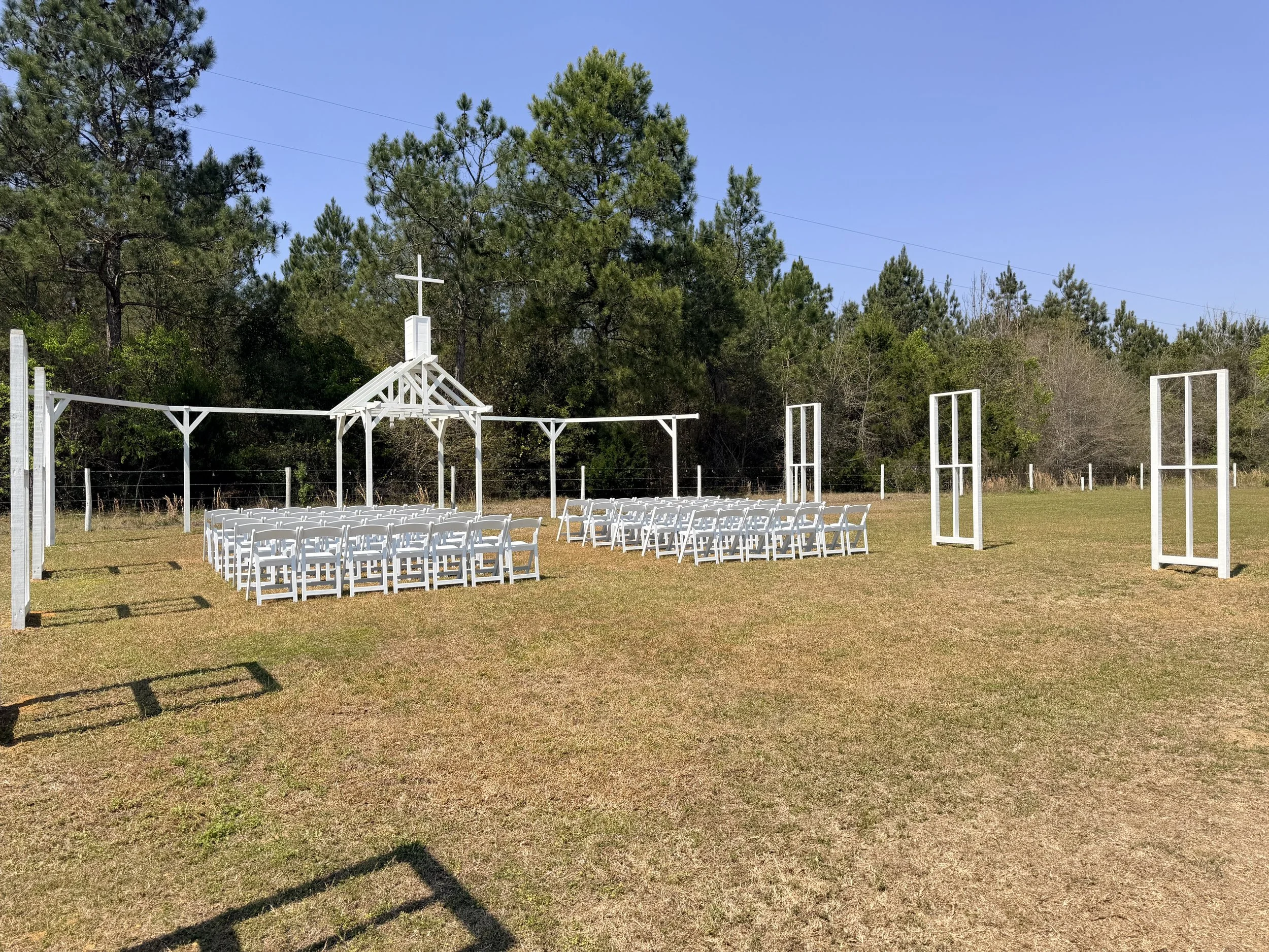 Open Air Chapel wedding ceremony setup with white chairs arranged in rows, white wooden frames as an aisle, and a small white chapel with a cross on top, set against a backdrop of trees and a clear blue sky.