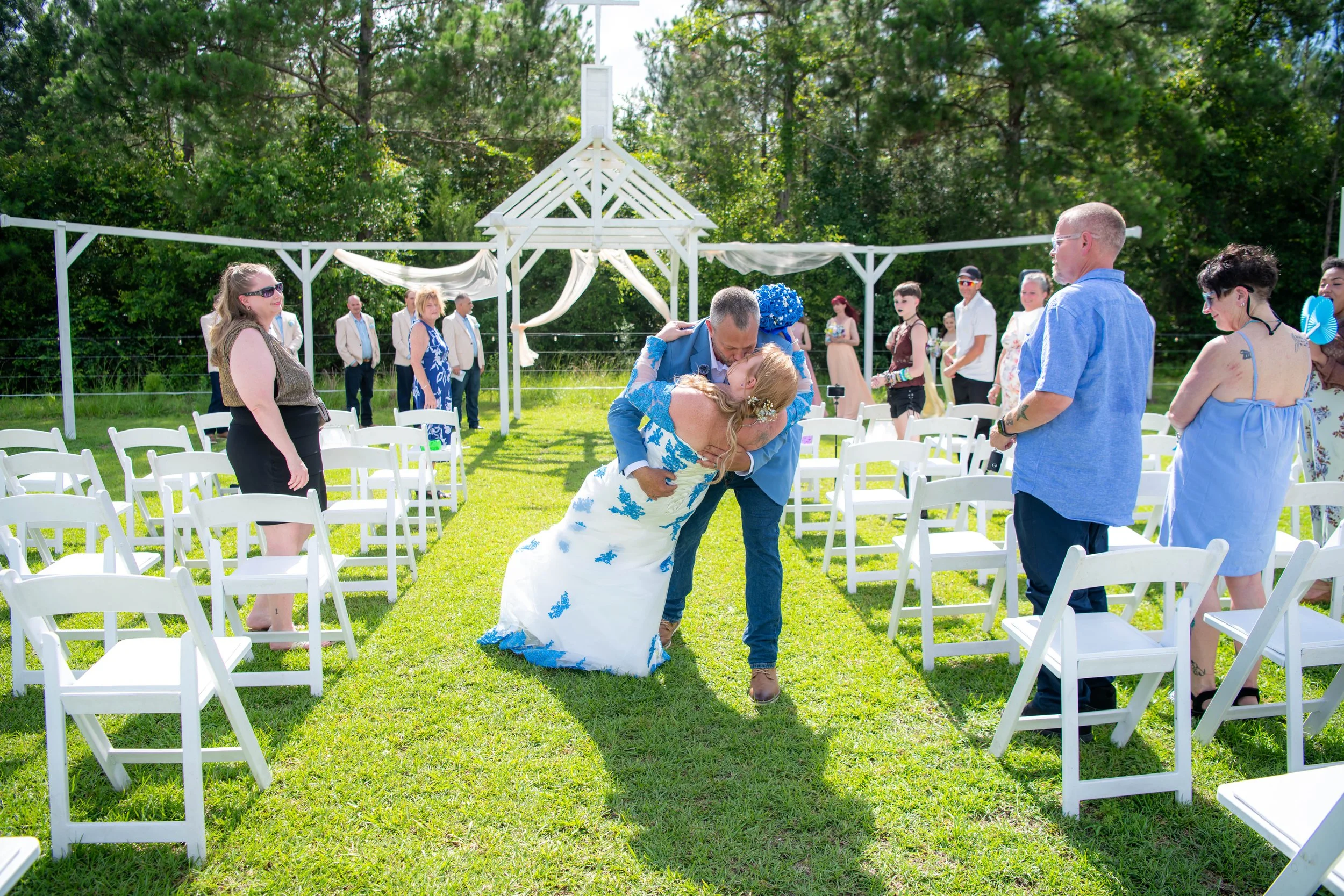 A wedding scene outdoors with the bride and groom dancing as the bride's father lifts her. Guests are seated and standing around the grassy area with white chairs and a white decorated archway in the background, surrounded by green trees.