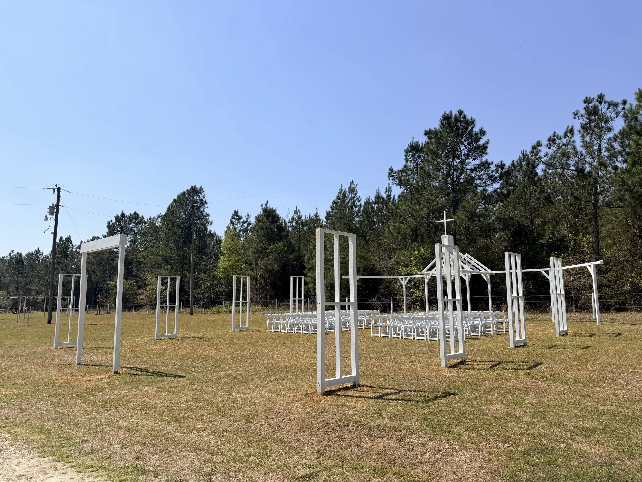An Open Air Chapel for a religious ceremony with white chairs arranged in a semi-circle and white wooden frames for a cross, all set on a grassy field with a background of pine trees and a blue sky.