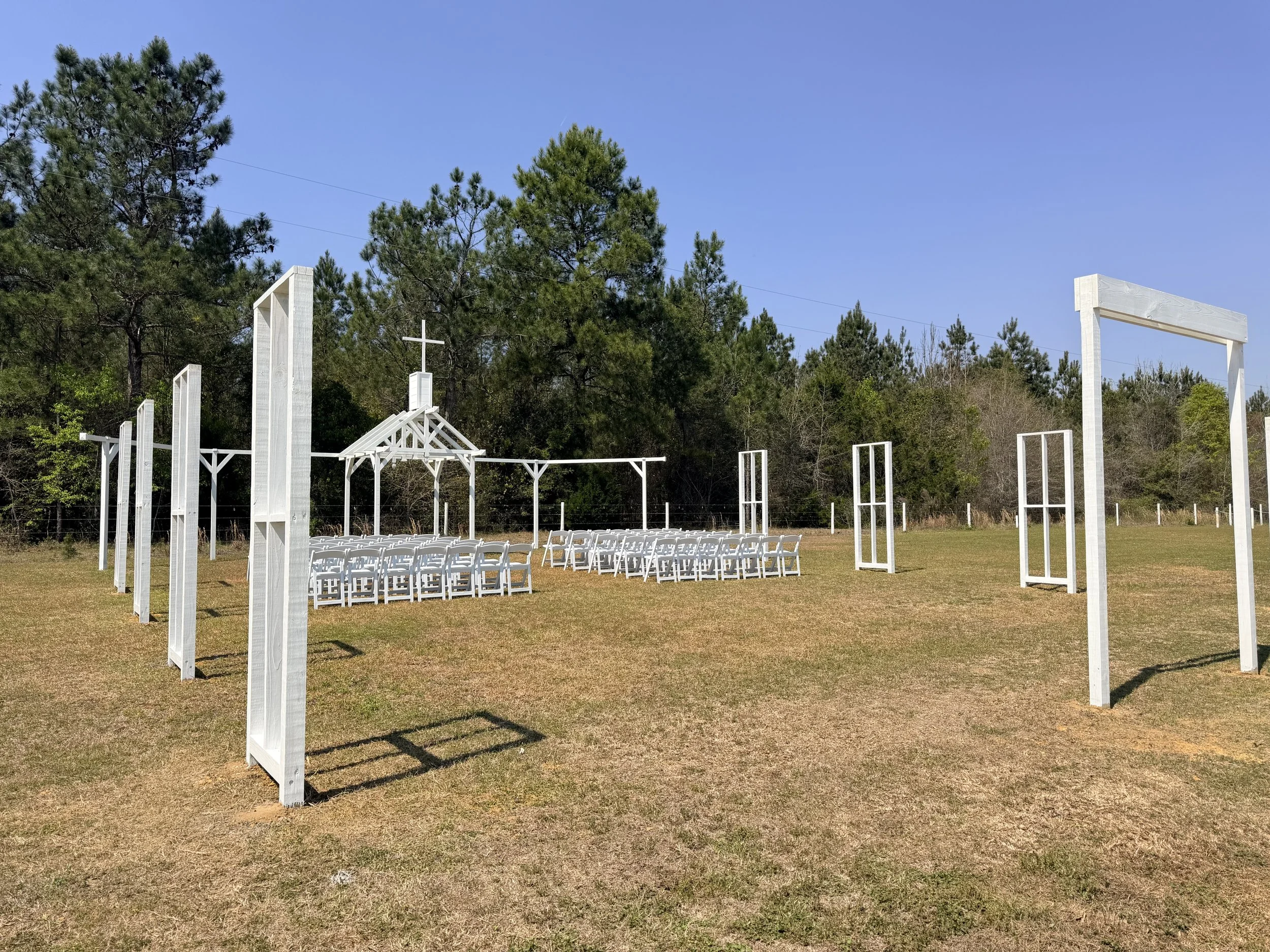 Open Air Chapel ceremony setup with white wooden arches, chairs, and a small chapel with a cross, on a grassy field surrounded by trees under a blue sky.