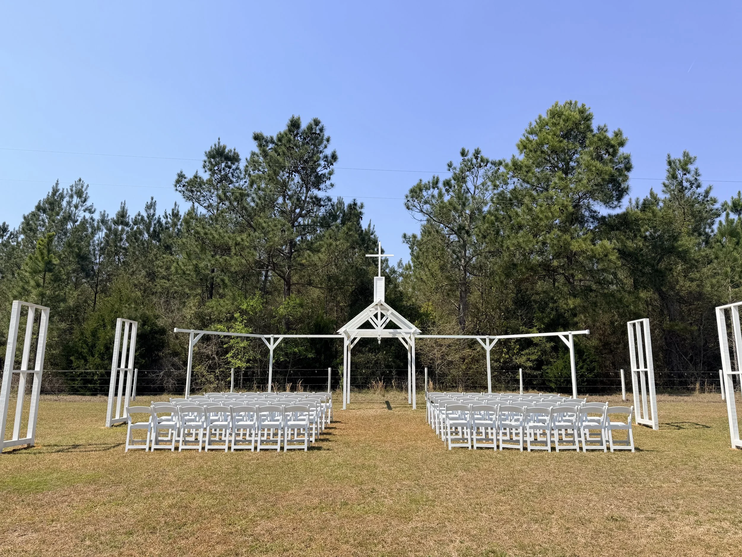Open Air Chapel wedding ceremony setup on grass with white chairs, arch, and open window frames under clear blue sky, surrounded by trees.