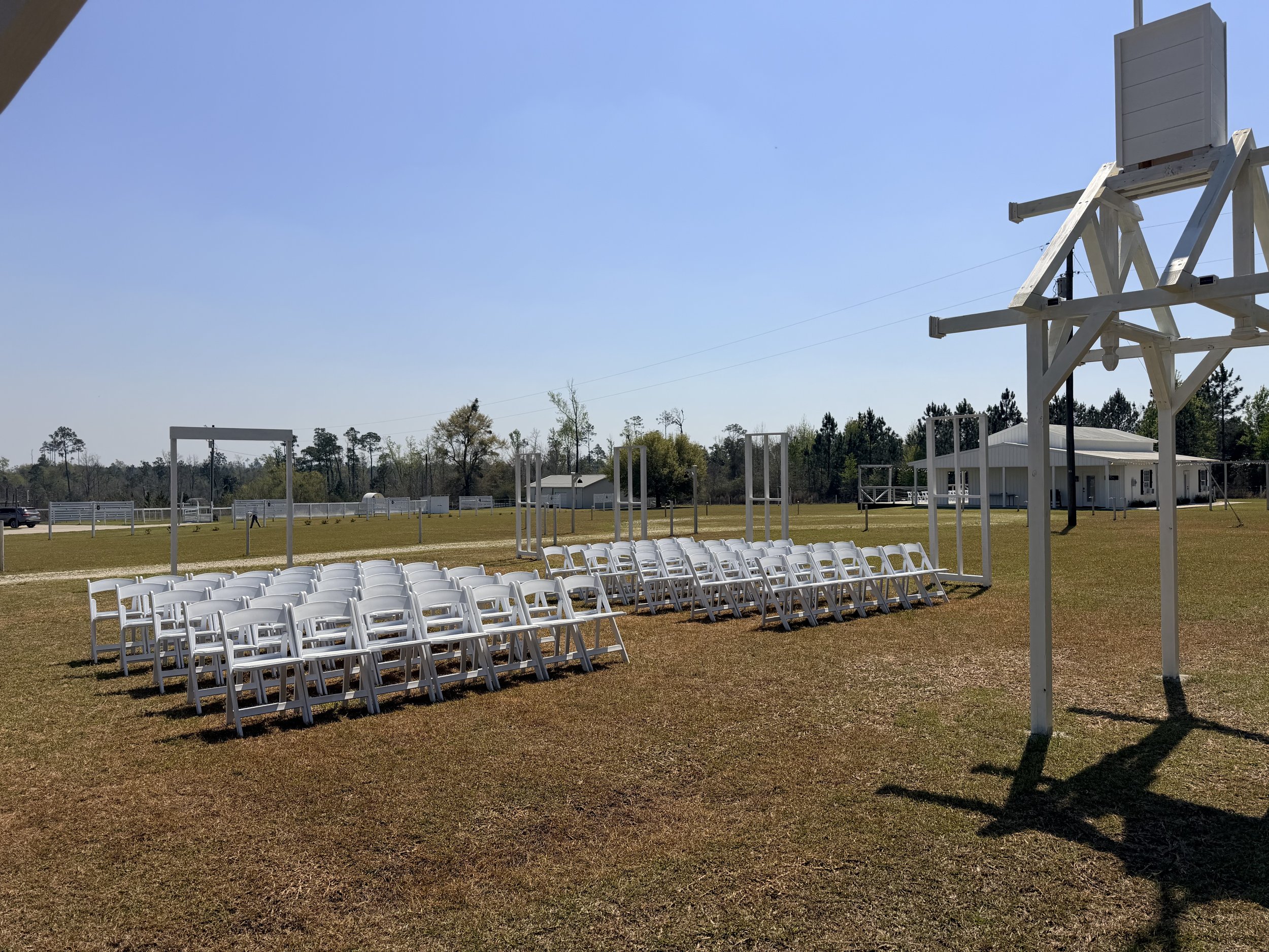 Open Air Chapel wedding or event setup with rows of white chairs and a small stage, on a grassy field under a clear blue sky.