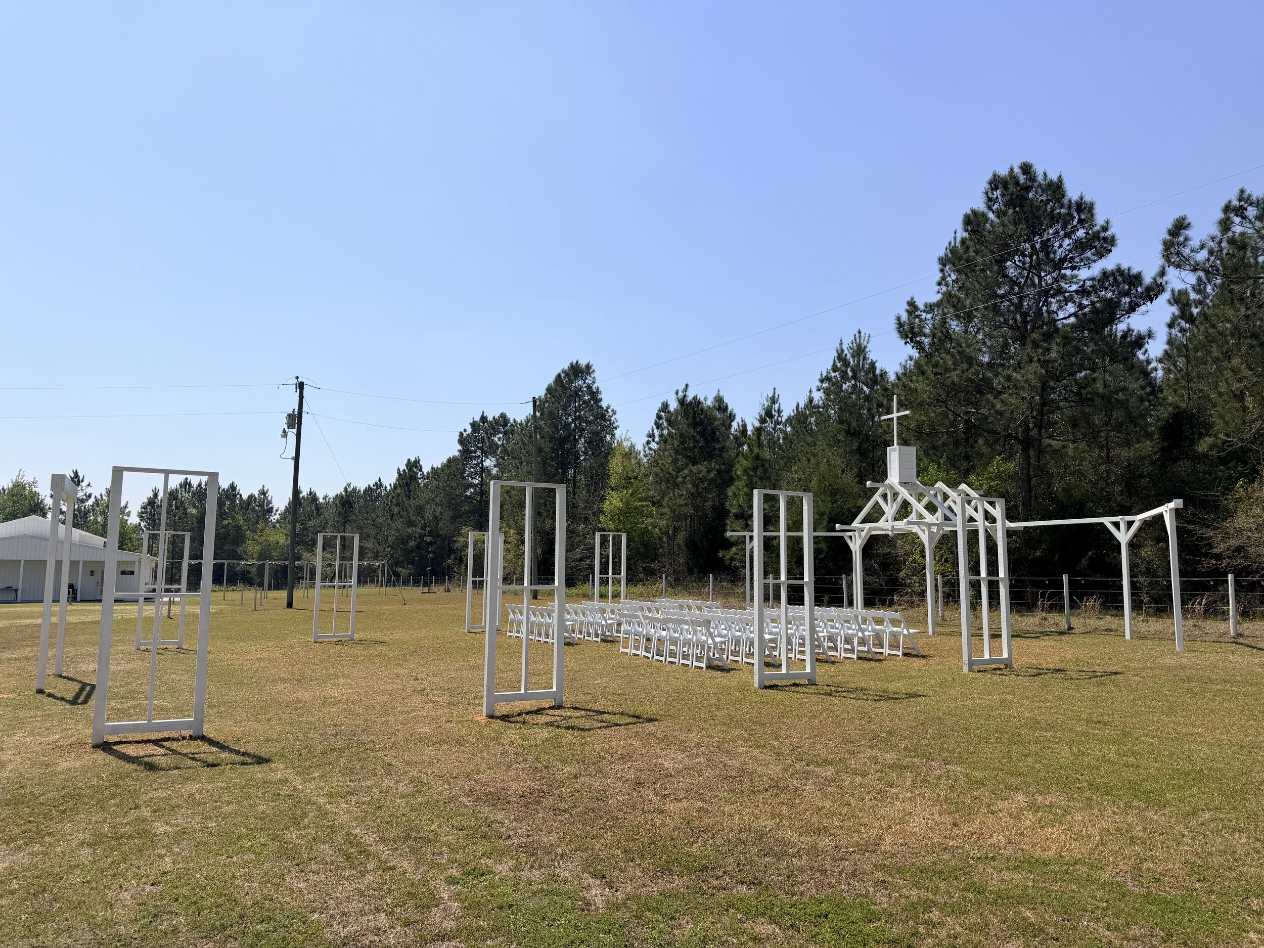 Open Air Chapel setup with white chairs, a cross, and a white pergola on a grassy field surrounded by trees.