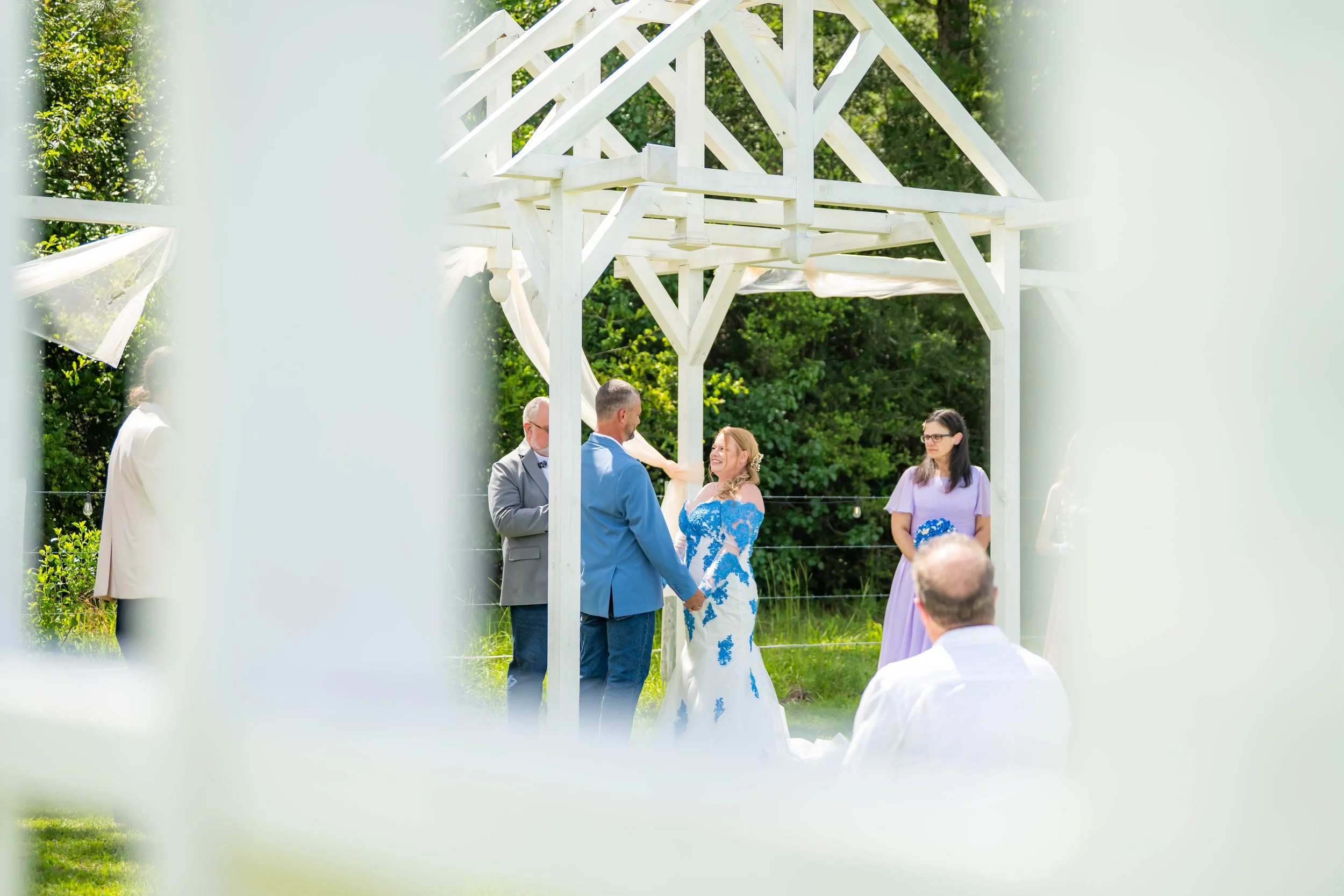A wedding ceremony outdoors, seen through decorative white fence posts, with a couple holding hands and exchanging vows under a white wooden arbor, surrounded by green trees and guests.