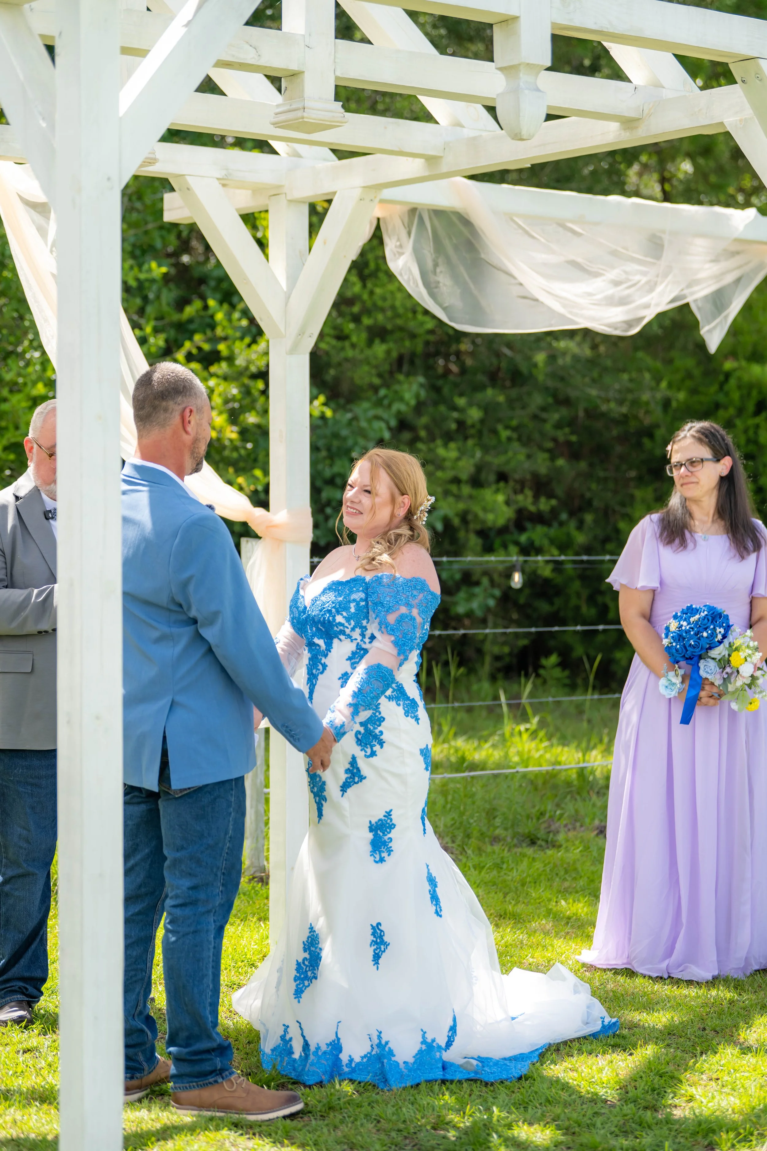 A wedding ceremony outdoors under a white wooden arbor with light fabric draping. The bride and groom are holding hands and exchanging vows, with the bride in a white gown with blue embroidery and the groom in a blue suit. A woman in a lavender dress
