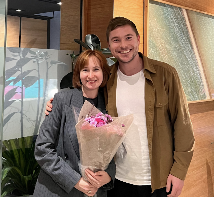 A smiling woman in a gray blazer holding a bouquet of pink flowers, standing next to a smiling man in a brown jacket and white t-shirt, in an indoor setting with plants and wooden decor.