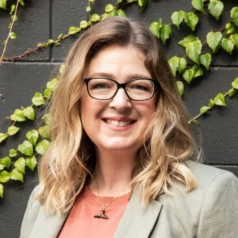 Taryn Sexton, smiling, wearing glasses, standing in front of a vine-covered wall.