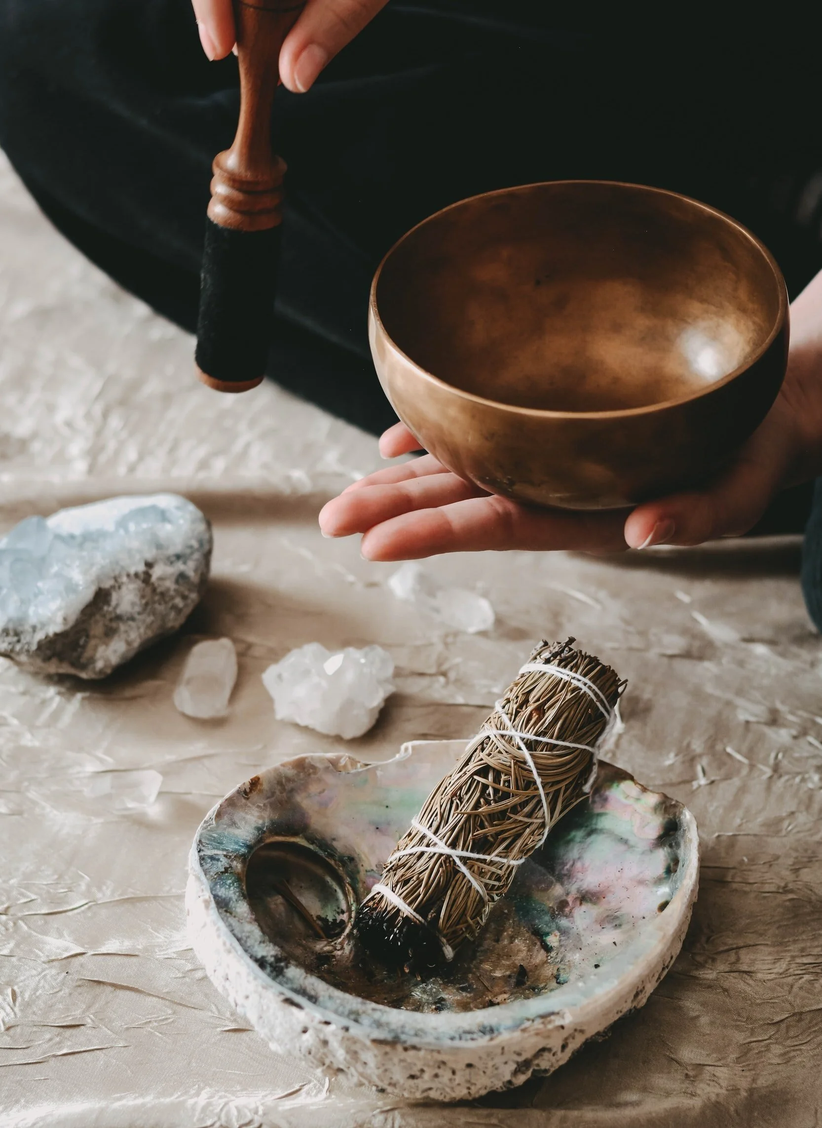 photo of hands holding a Tibetan singing bowl over a dried bundle of rosemary in an abalone shell alongside crystals