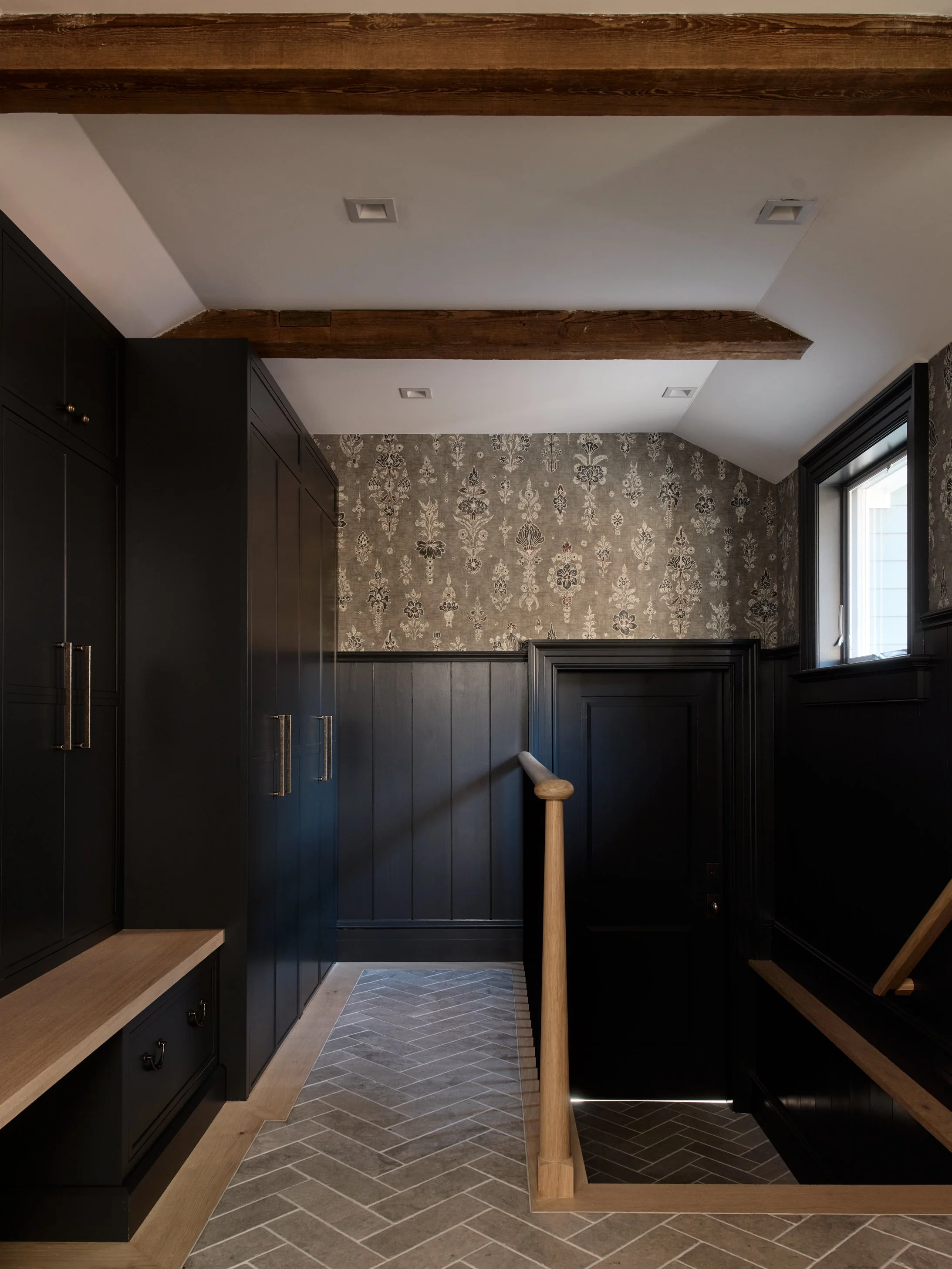 Interior of a room with dark cabinetry, a patterned wallpapered wall, a large window, and a tiled floor with a herringbone pattern. A wooden railing is visible.