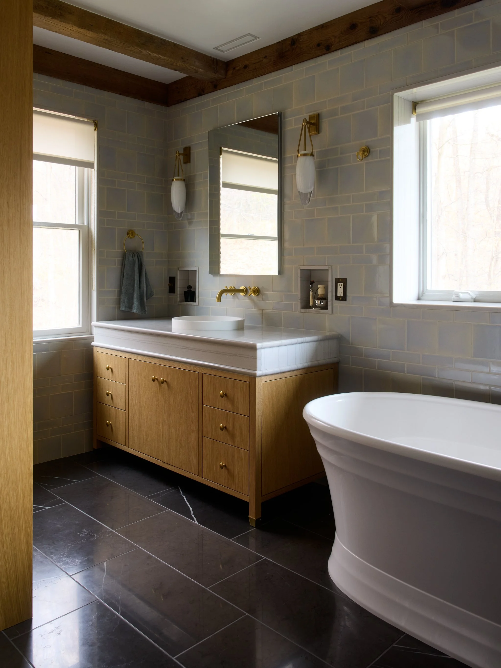 A bathroom with dark tiled floor, wooden cabinetry, a white freestanding bathtub, a large window, and a mirror above the sink with gold fixtures.