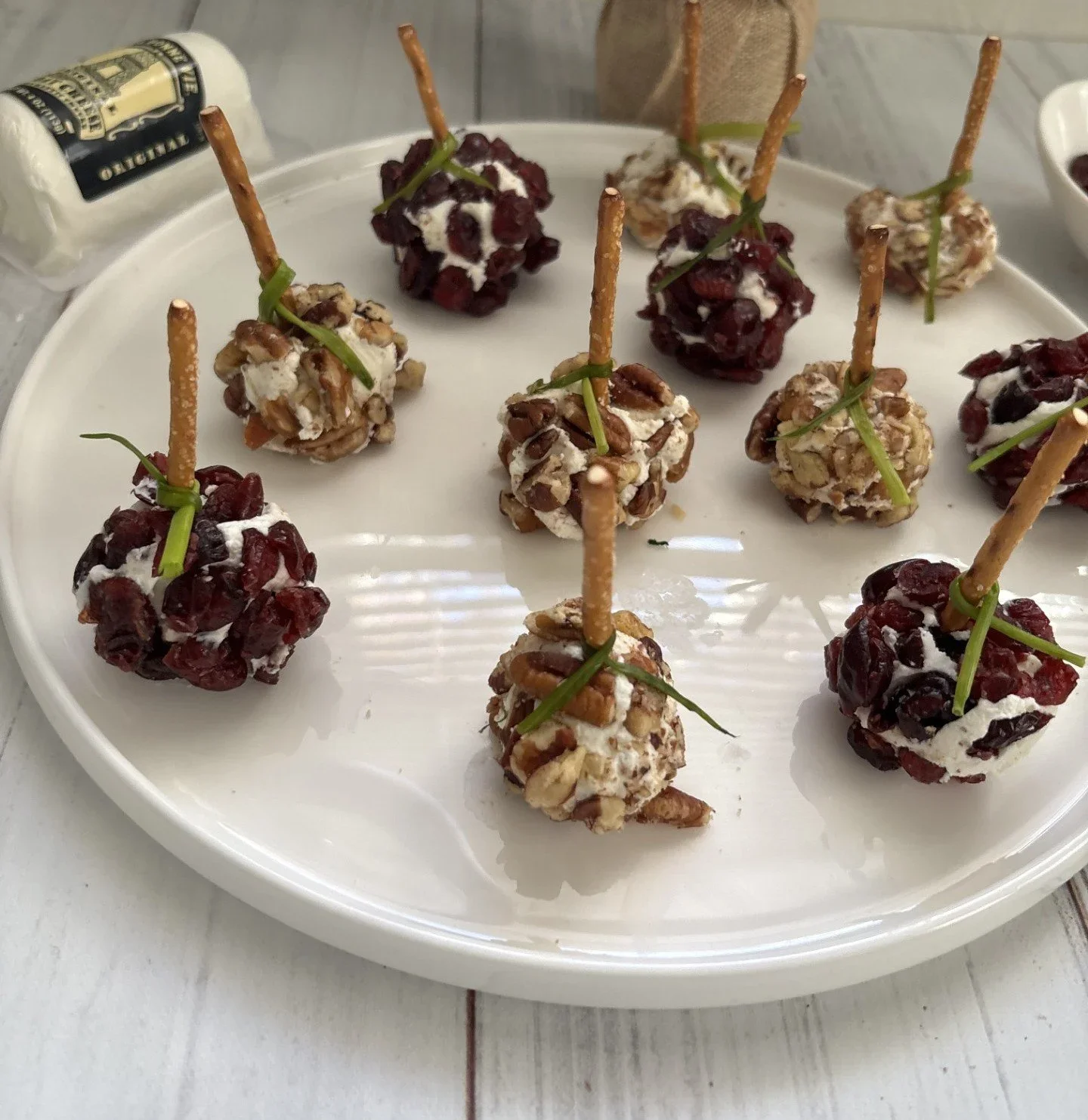 Bite-sized balls covered in nuts and dried cranberries, garnished with thin green herbs, served on a white platter.