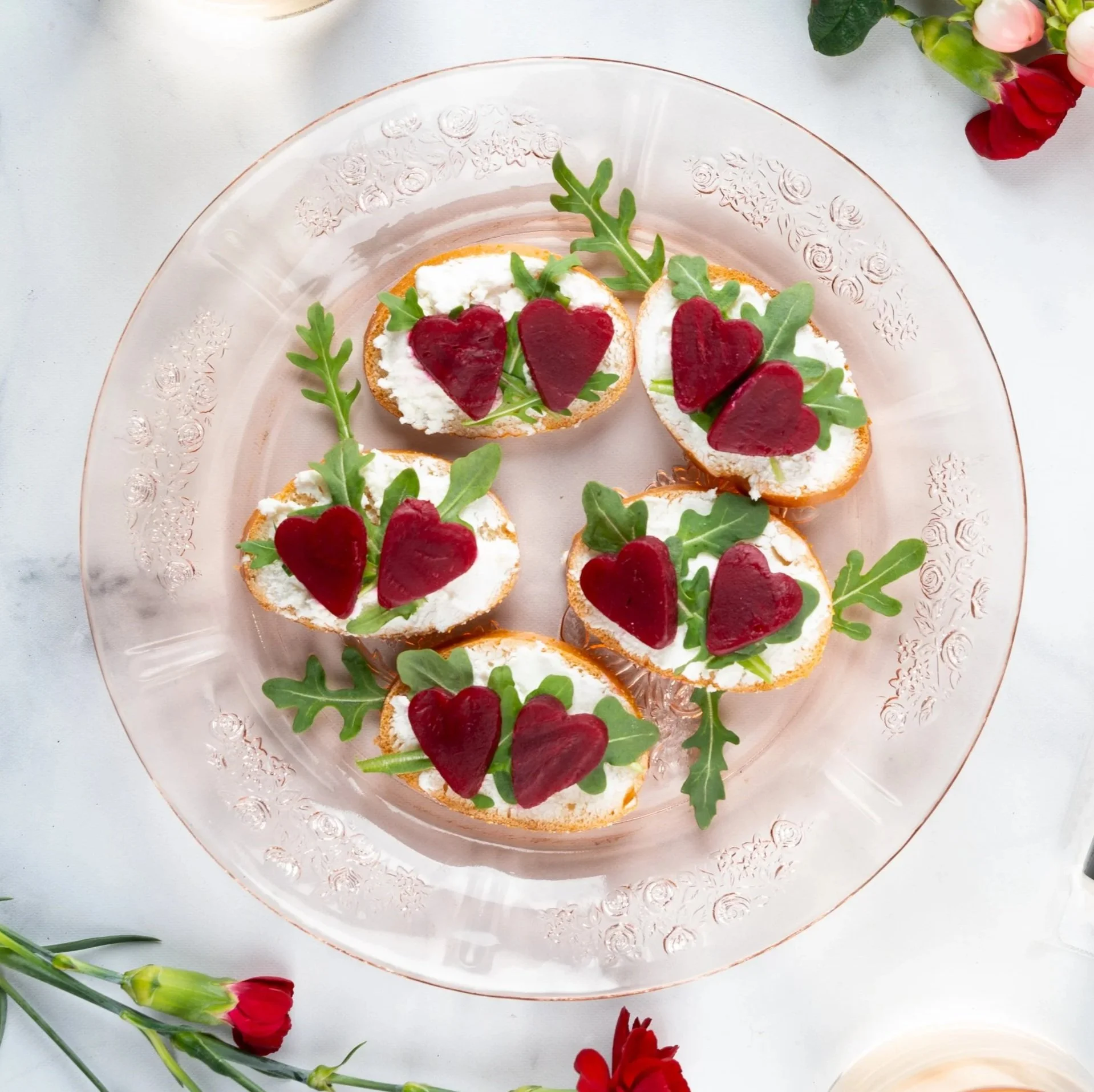 Canapés with cream cheese, red heart-shaped gelatin, and arugula on a glass plate.
