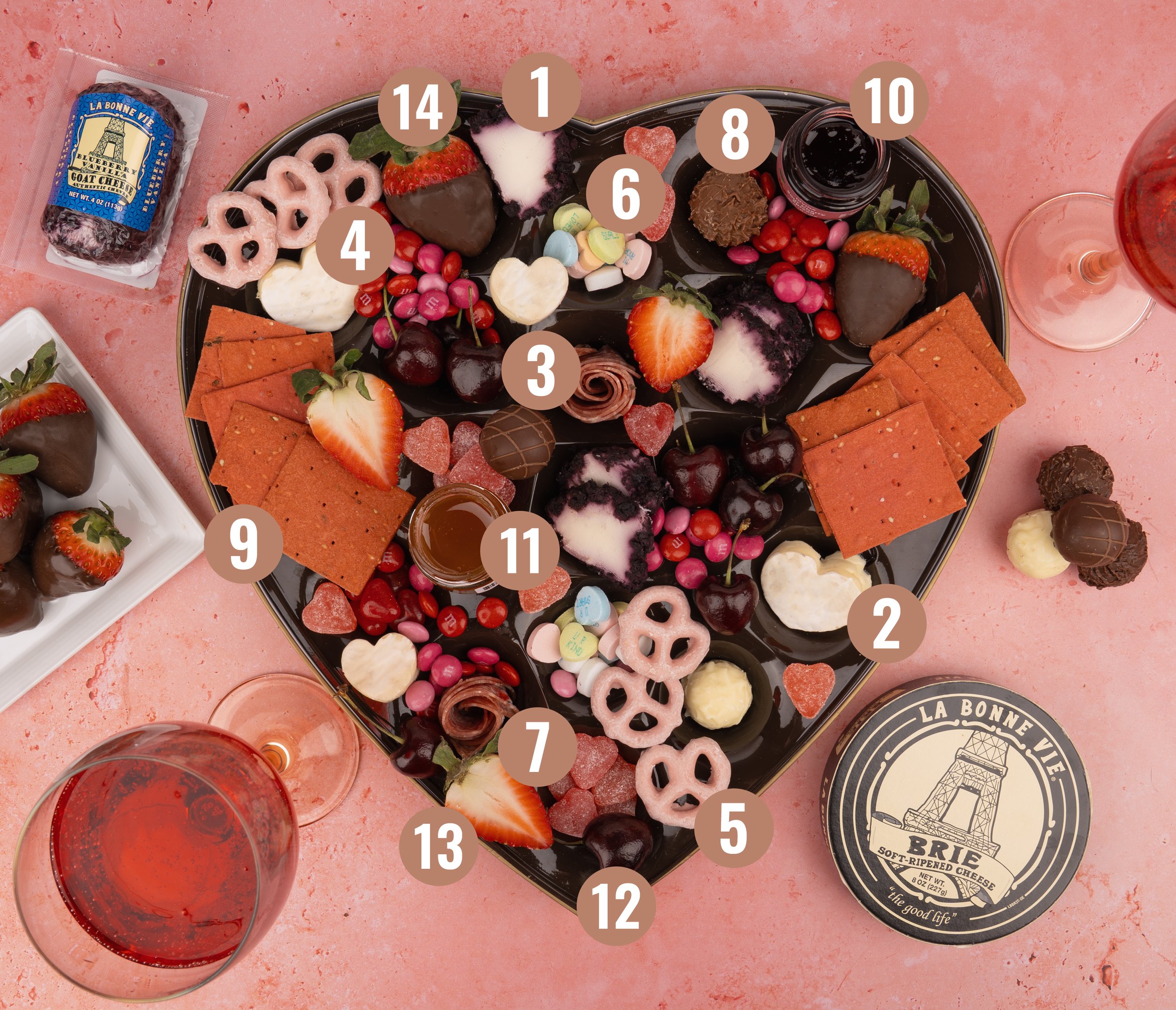 Heart-shaped chocolate box with La Bonne Vie Brie and Blueberry Vanilla goat cheese with an assortment of chocolates, strawberries, candies, crackers, honey, and chocolates, surrounded by wine glasses on a pink surface.