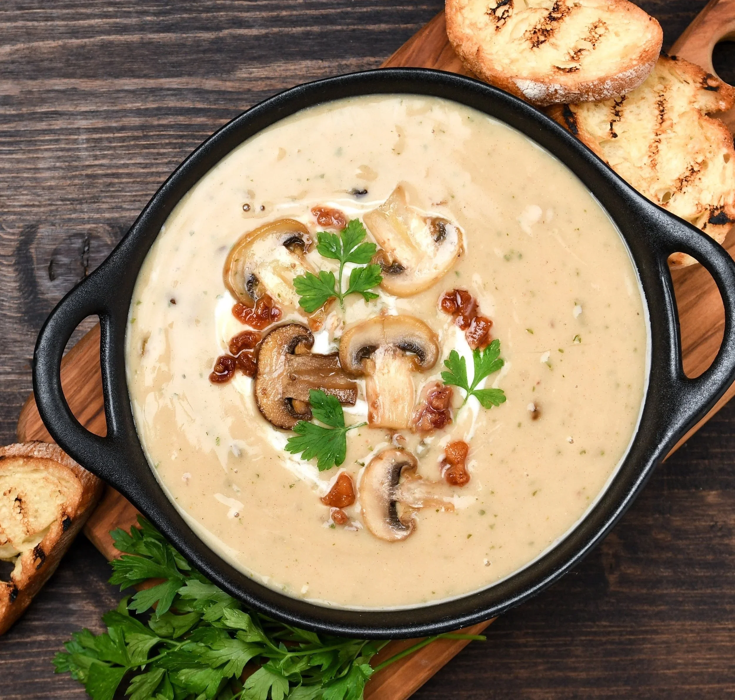 Mushroom and herb creamy soup garnished with parsley served in a black bowl on a wooden board, with toasted bread slices nearby.