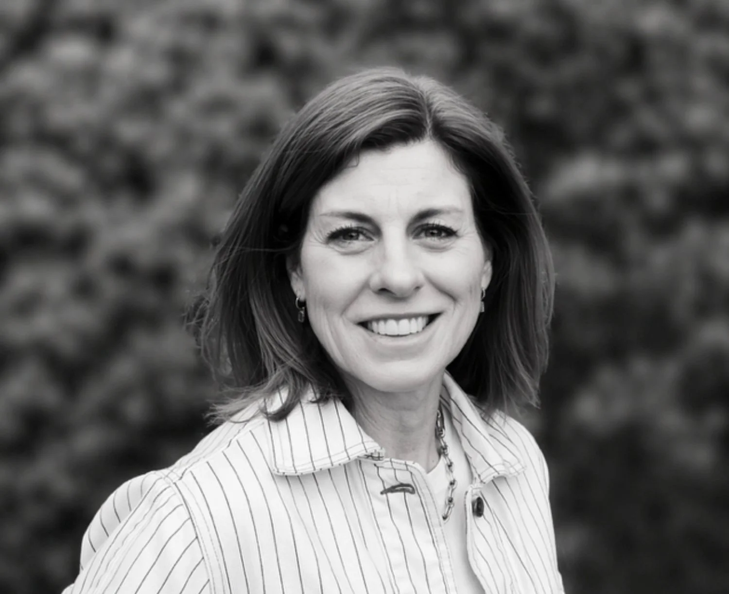 Black and white close up portrait of smiling studio manager Jo Chambers wearing a striped jacket against a leafy background