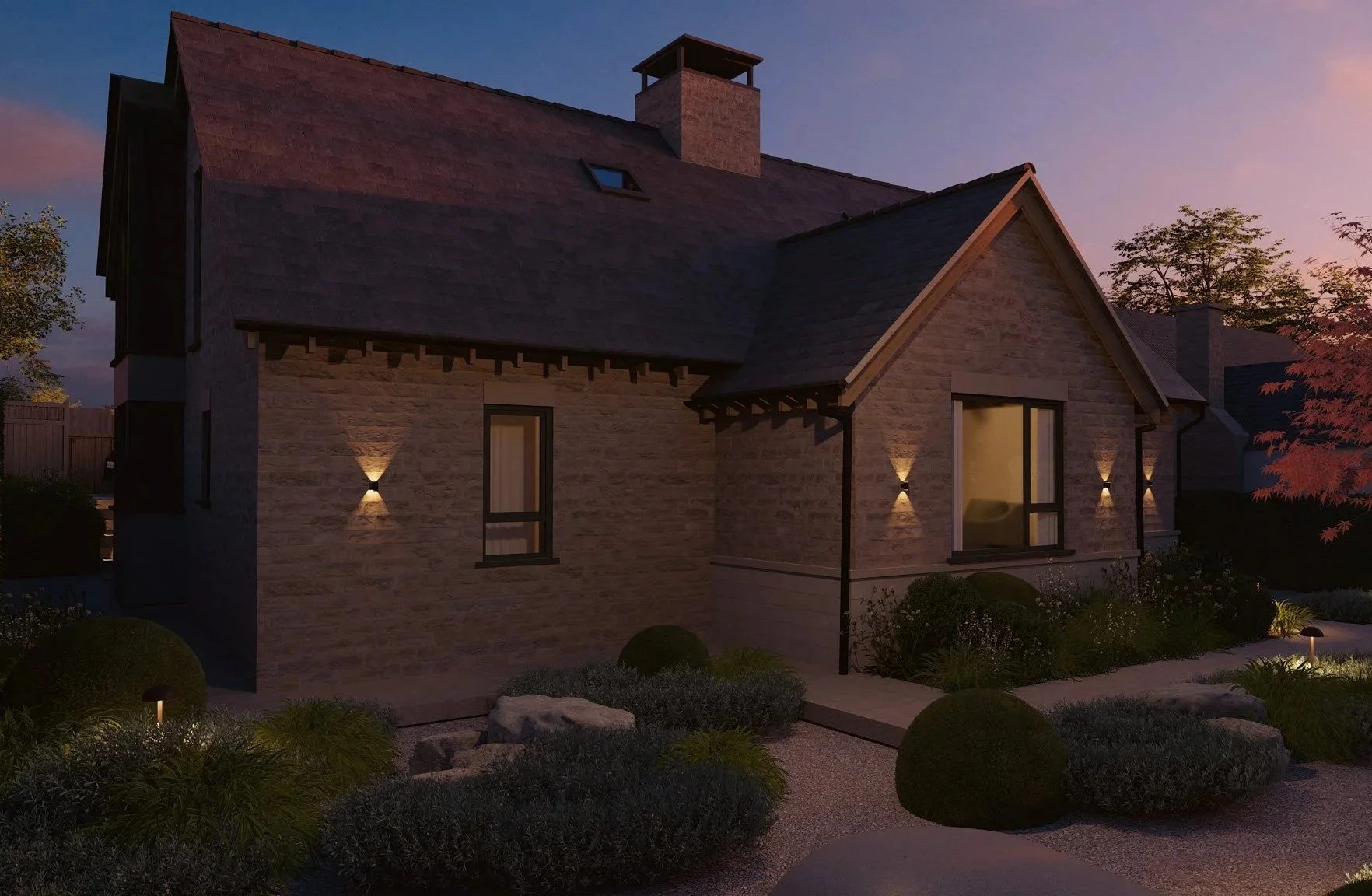 A two-story house with stone exterior walls, illuminated by exterior wall lights, during dusk or twilight. The house has a steep sloped roof, a chimney, windows, and a small landscaped yard with bushes, rocks, and pathway lighting.