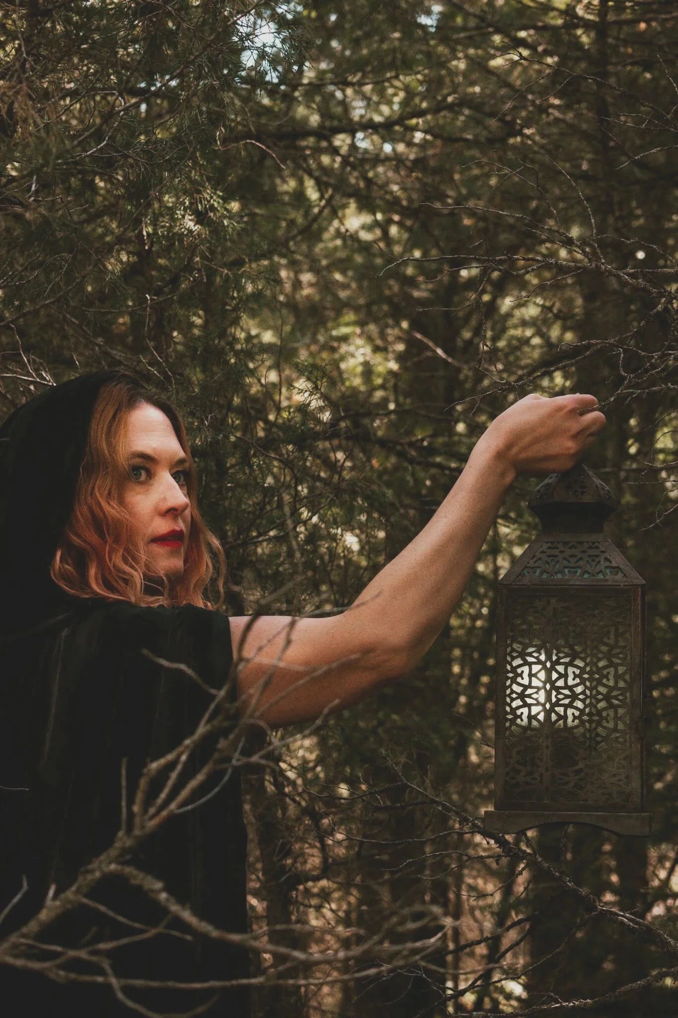 A woman with red wavy hair, wearing a black hooded cloak, touches a decorative lantern hanging from a tree in a forest setting.