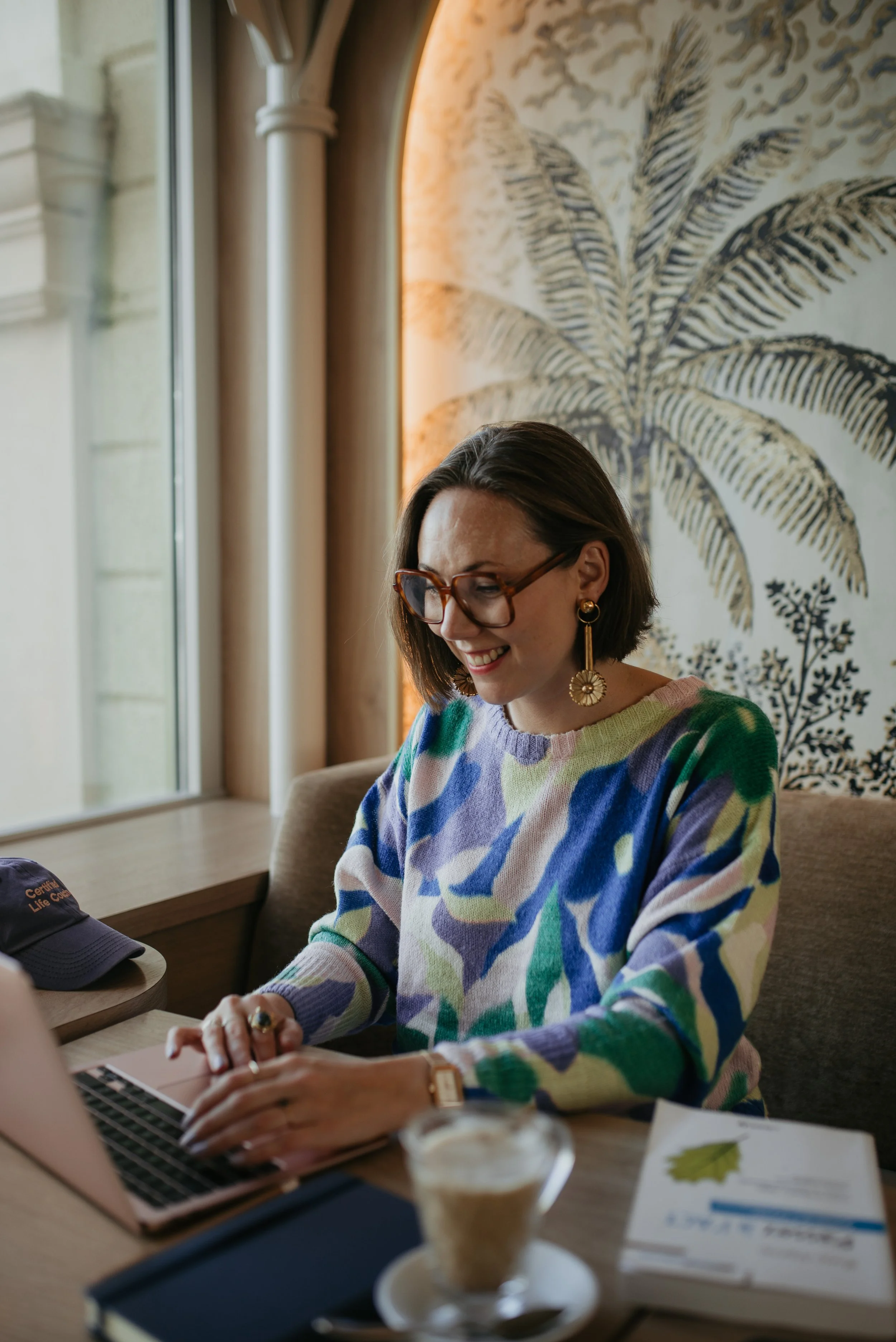 A woman with glasses and earrings working on a laptop in a cozy cafe with a palm tree mural on the wall.