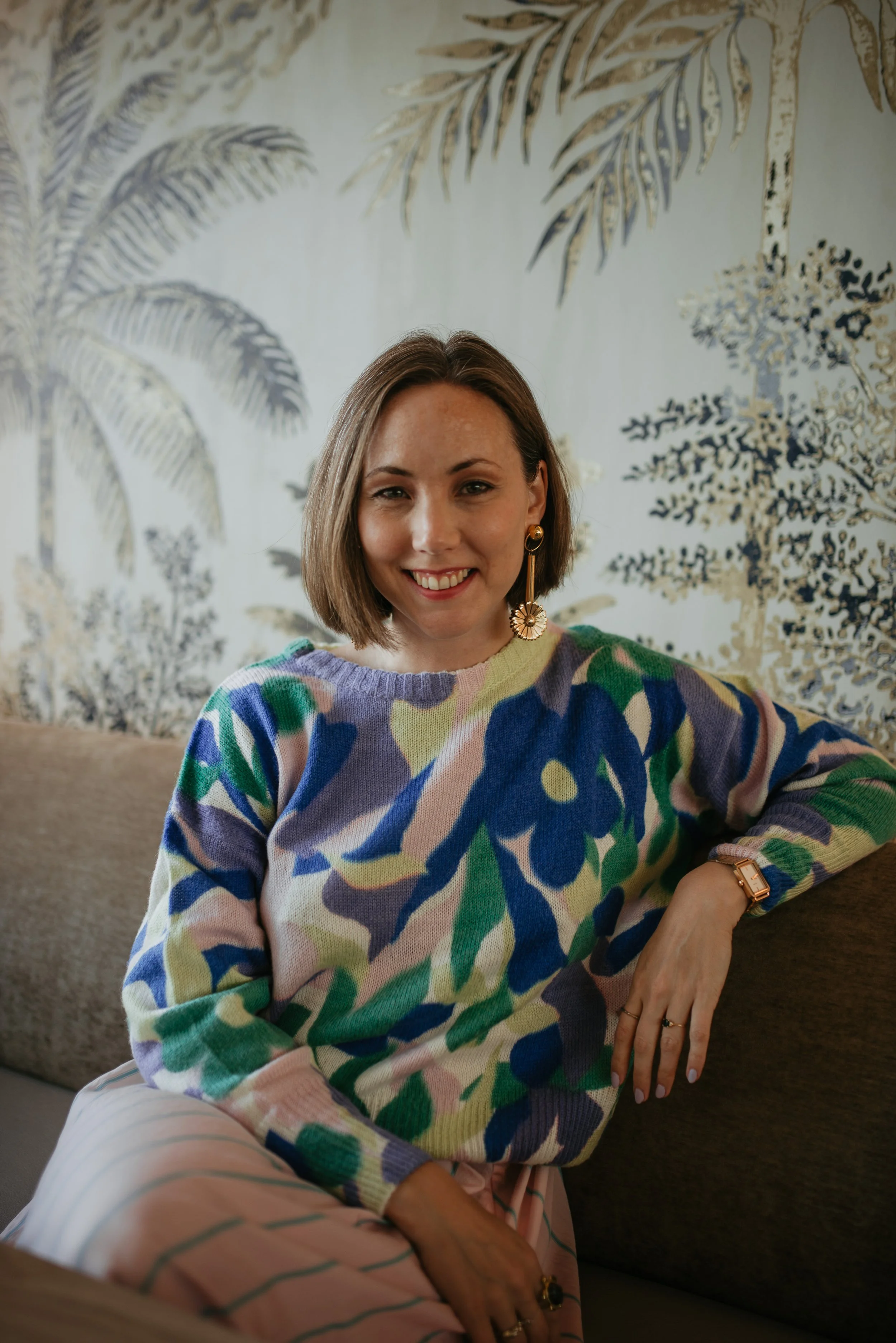 A woman sitting on a sofa, smiling at the camera, in a room with a decorative wall featuring palm tree artwork, wearing a colorful patterned sweater, a watch, earrings, and rings.