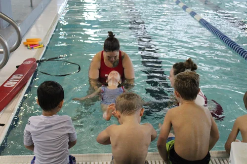Lifeguard supports young swimmer who is learning to float on her back while other children sit on the side of the pool and watch