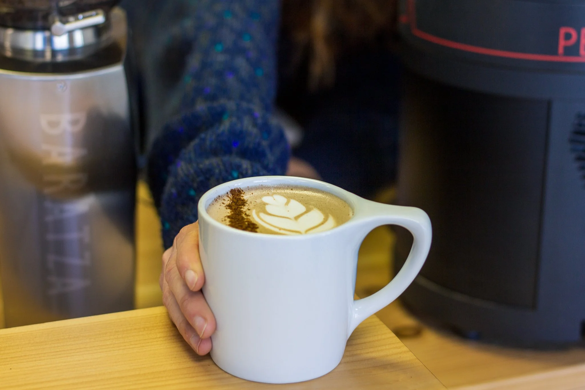 A hand holding a white mug with latte art on coffee, in a café setting.