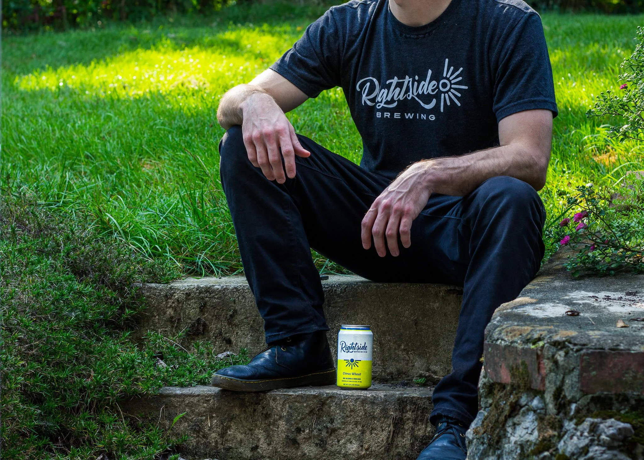 A man sitting outdoors on concrete steps, wearing a black T-shirt that says 'Rightside Brewing' and black pants, with a can of Rightside Brewing Citrus Wheat beer placed beside him on the steps.