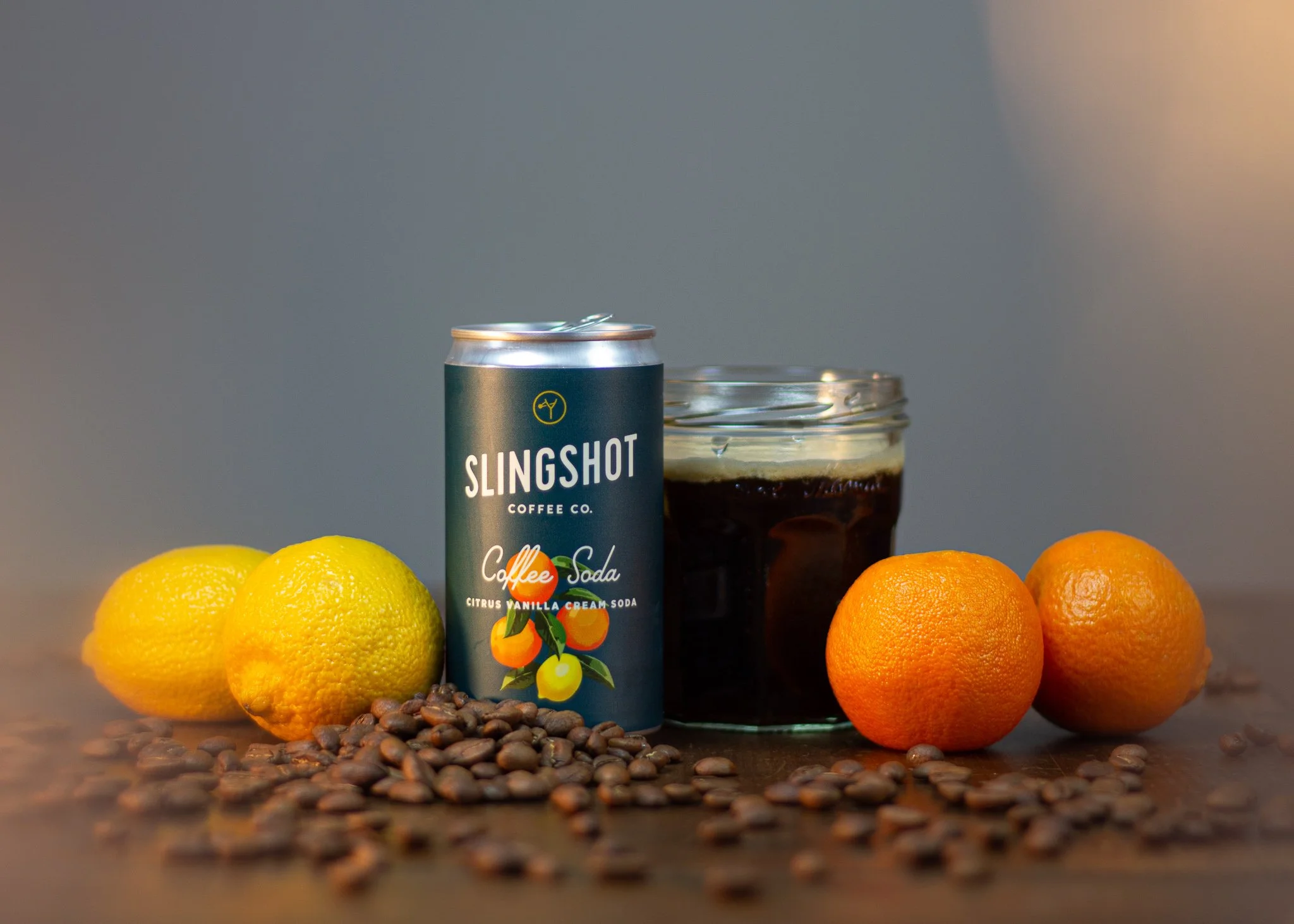 A can of Slingshot Coffee Co. citrus vanilla cream soda, a glass of dark soda, three lemons, two oranges, and scattered coffee beans on a wooden surface.