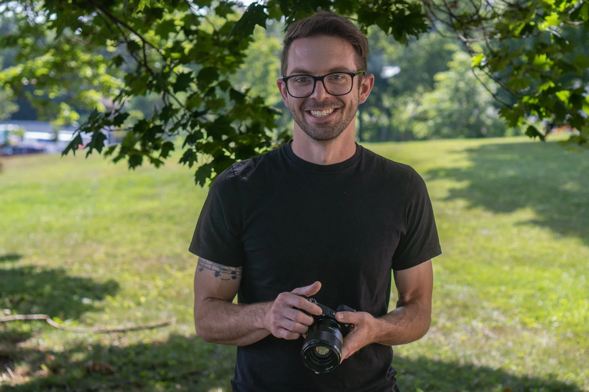 A smiling man with glasses and a black T-shirt standing outdoors under a tree, holding a camera.